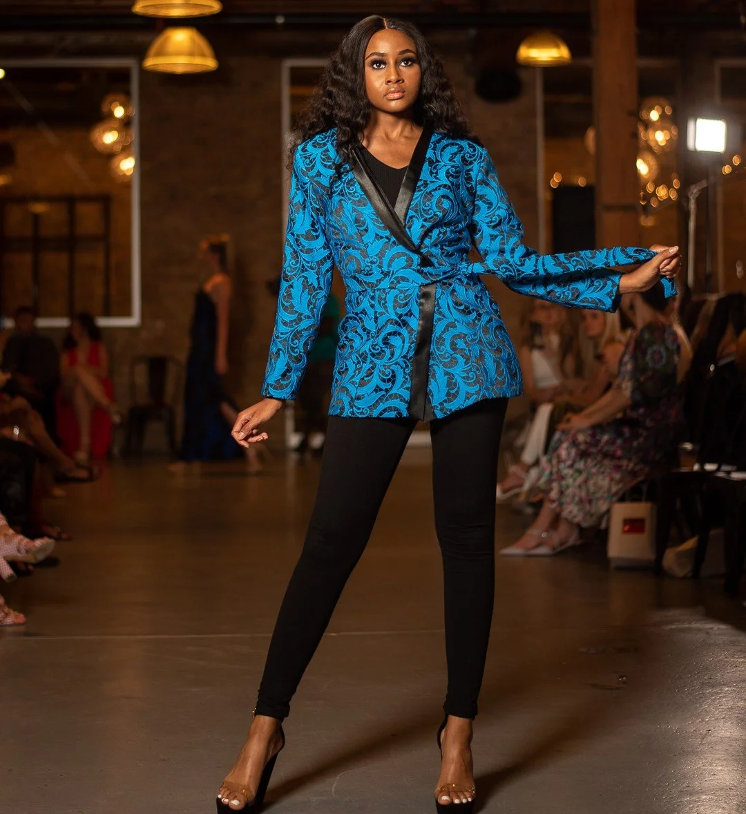 A woman modeling a blue and black embroidered blazer with black pants and high heels on a runway.