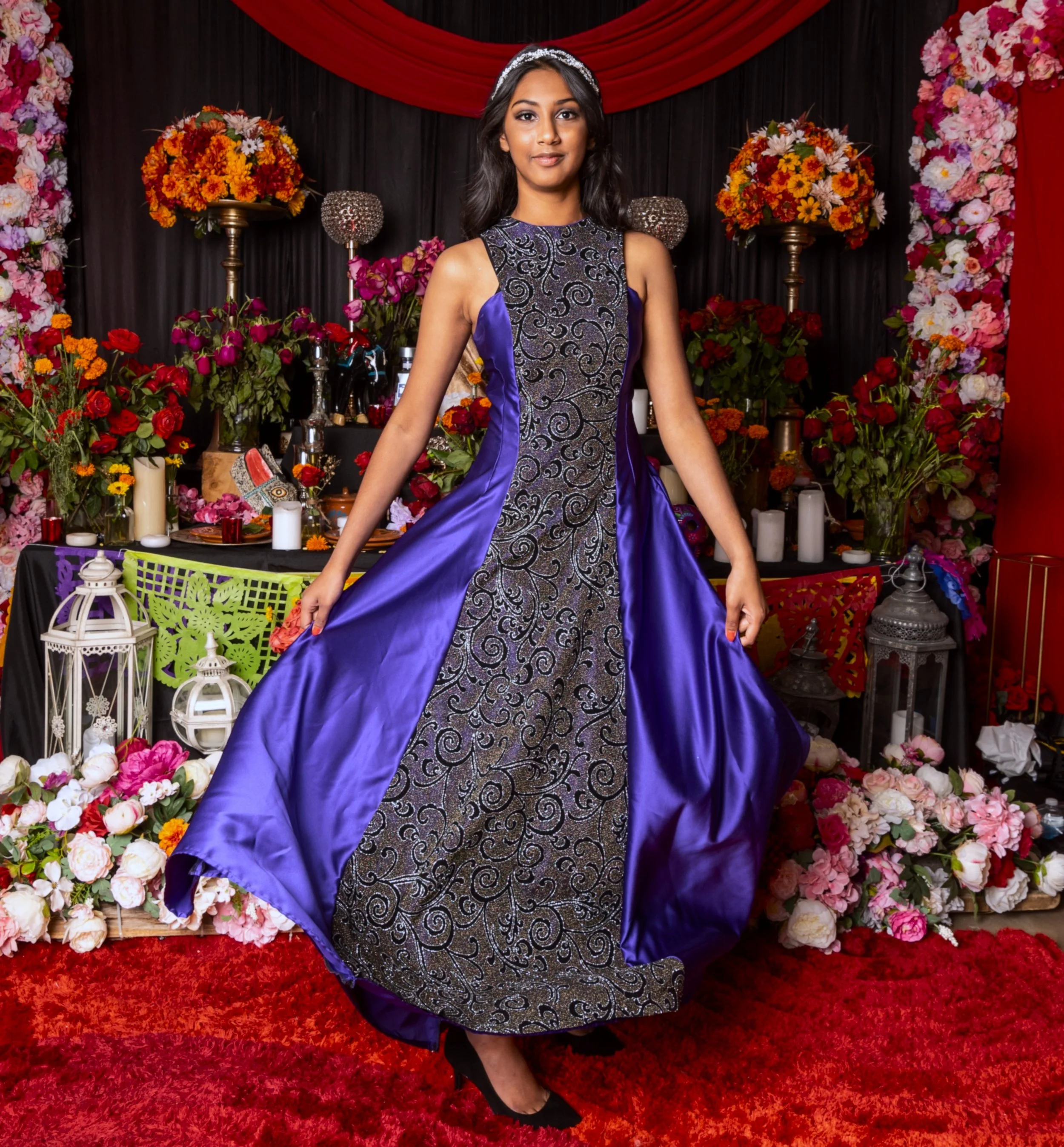 A woman in an elegant purple and black sleeveless gown stands in front of a floral and candlelit backdrop.