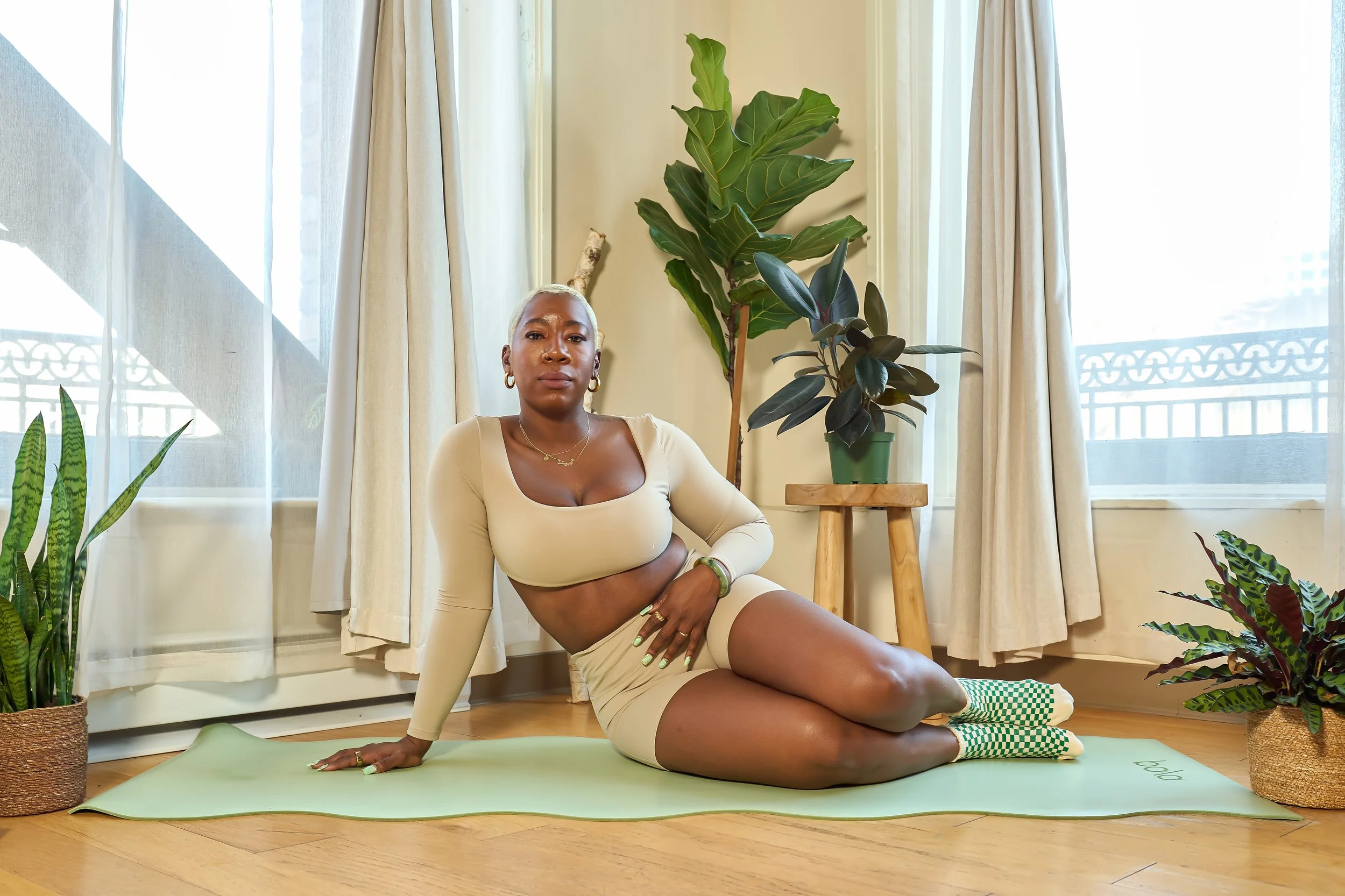 A woman with short blonde hair and dark skin sitting on a yoga mat during the daytime, surrounded by potted plants, in front of a large window with white curtains.
