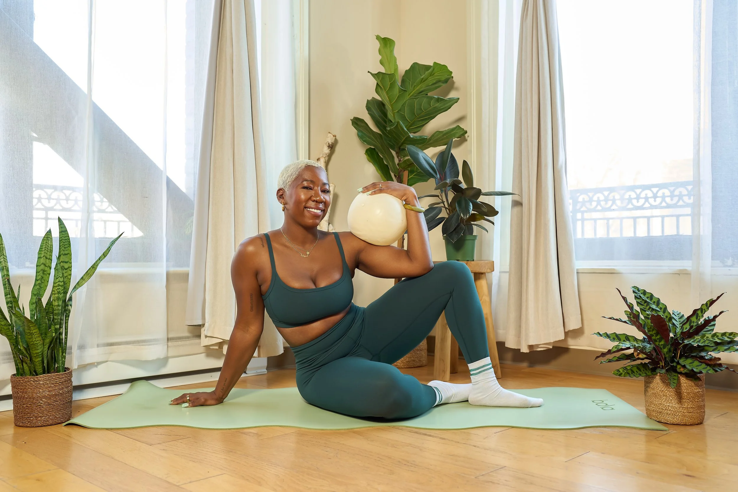 A woman with short blonde hair doing pilates indoors on a green mat, surrounded by houseplants, holding a ball on her shoulder, smiling, wearing a green sports bra, matching leggings, and white socks.