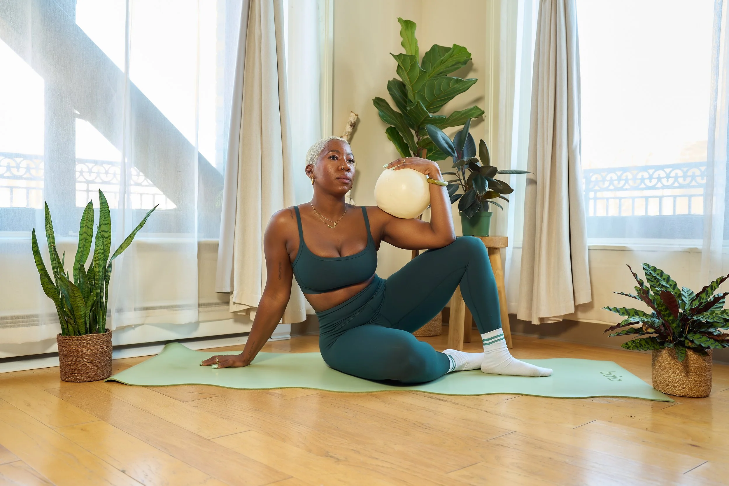 A woman with short platinum blonde hair practicing yoga indoors on a green mat surrounded by potted houseplants, with sunlight coming through sheer curtains in the background.