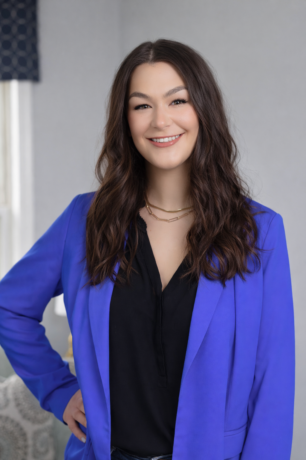 A woman with long wavy brown hair, smiling, wearing a blue blazer over a black shirt, and gold necklaces, standing indoors against a plain wall.