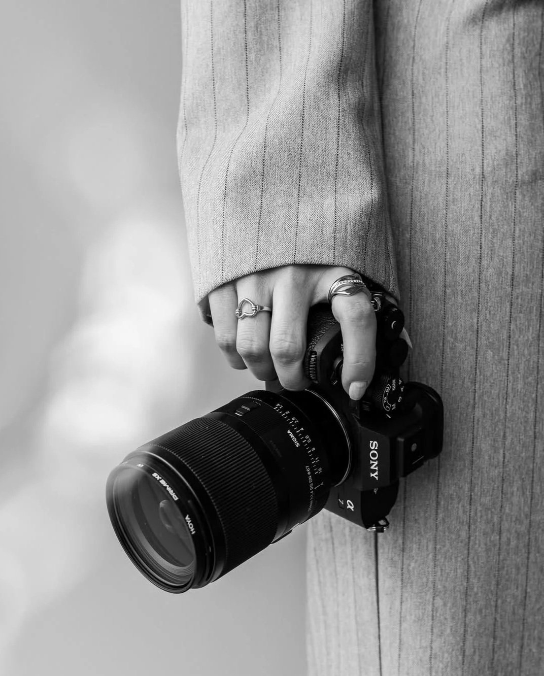 A person wearing a striped suit holding a Sony camera with a large lens in their right hand, with rings on their fingers.