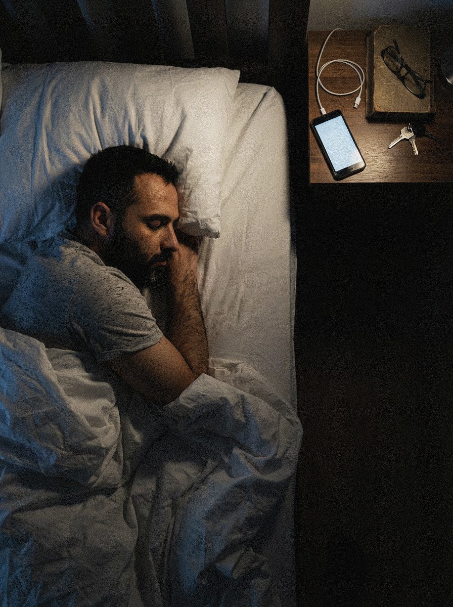 A man with dark hair and beard sleeping on his side in a bed with white sheets, next to a nightstand with a smartphone, a pair of glasses, keys, and an over-the-ear headset.