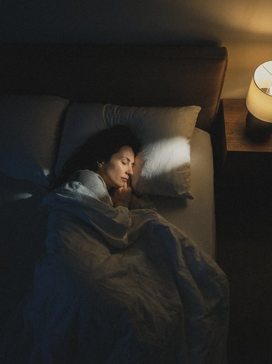 A woman sleeping in bed with a pillow, under a blanket, in a dimly lit room with a bedside lamp.
