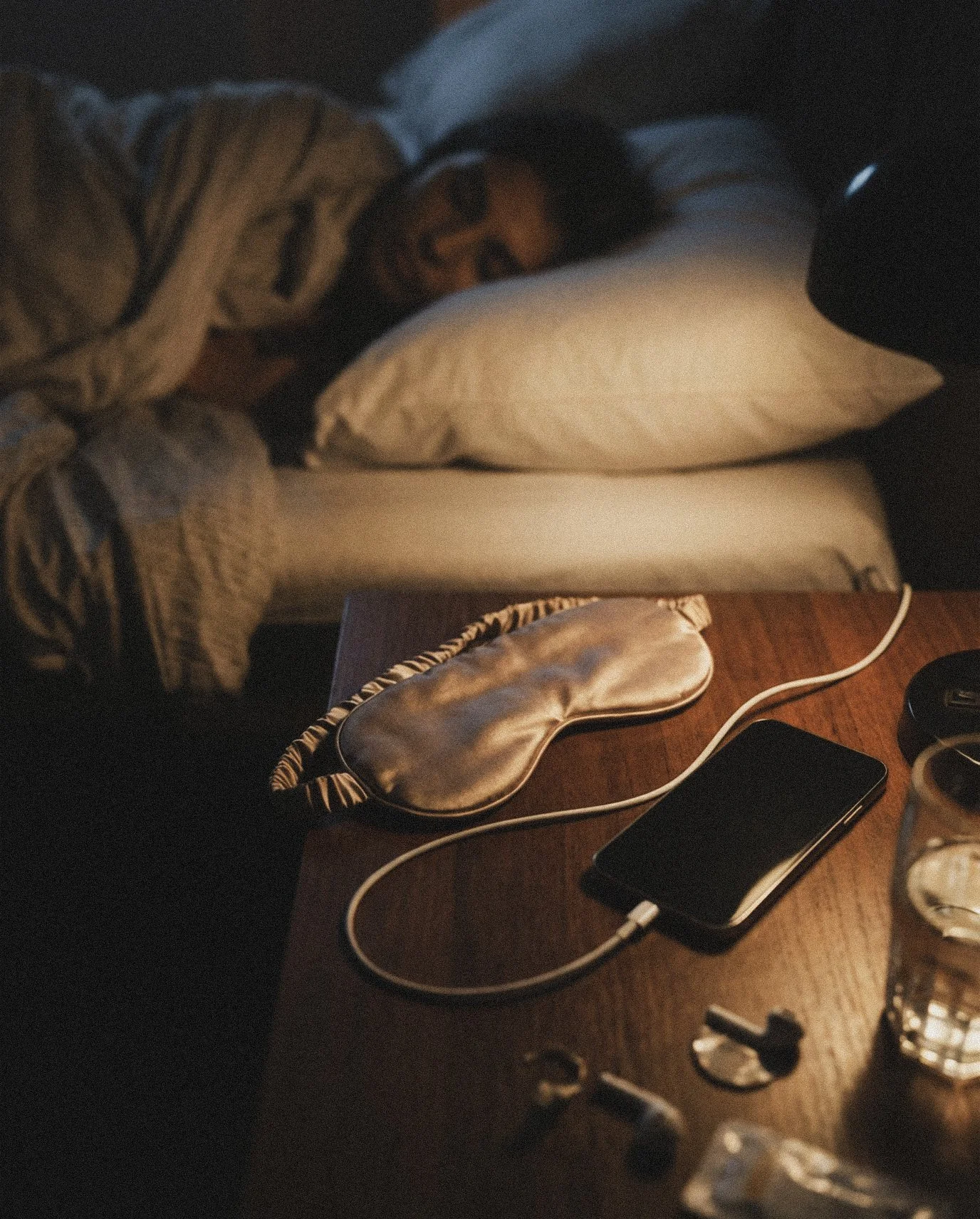 A woman lying on a bed with a pillow, wearing pajamas, illuminated by soft light. On a bedside table, there is a sleep mask, a smartphone, a pair of earbuds, a glass of water, and some small items.