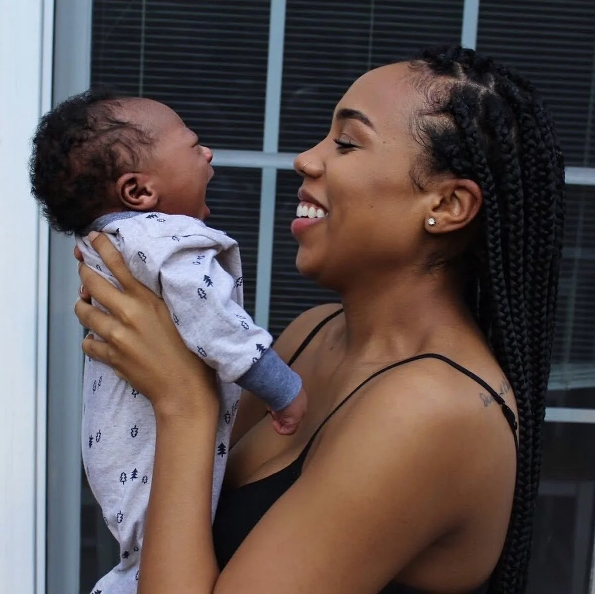A smiling woman with braided hair holds a baby near her face, both smiling and facing each other, outside in front of a window with closed blinds.