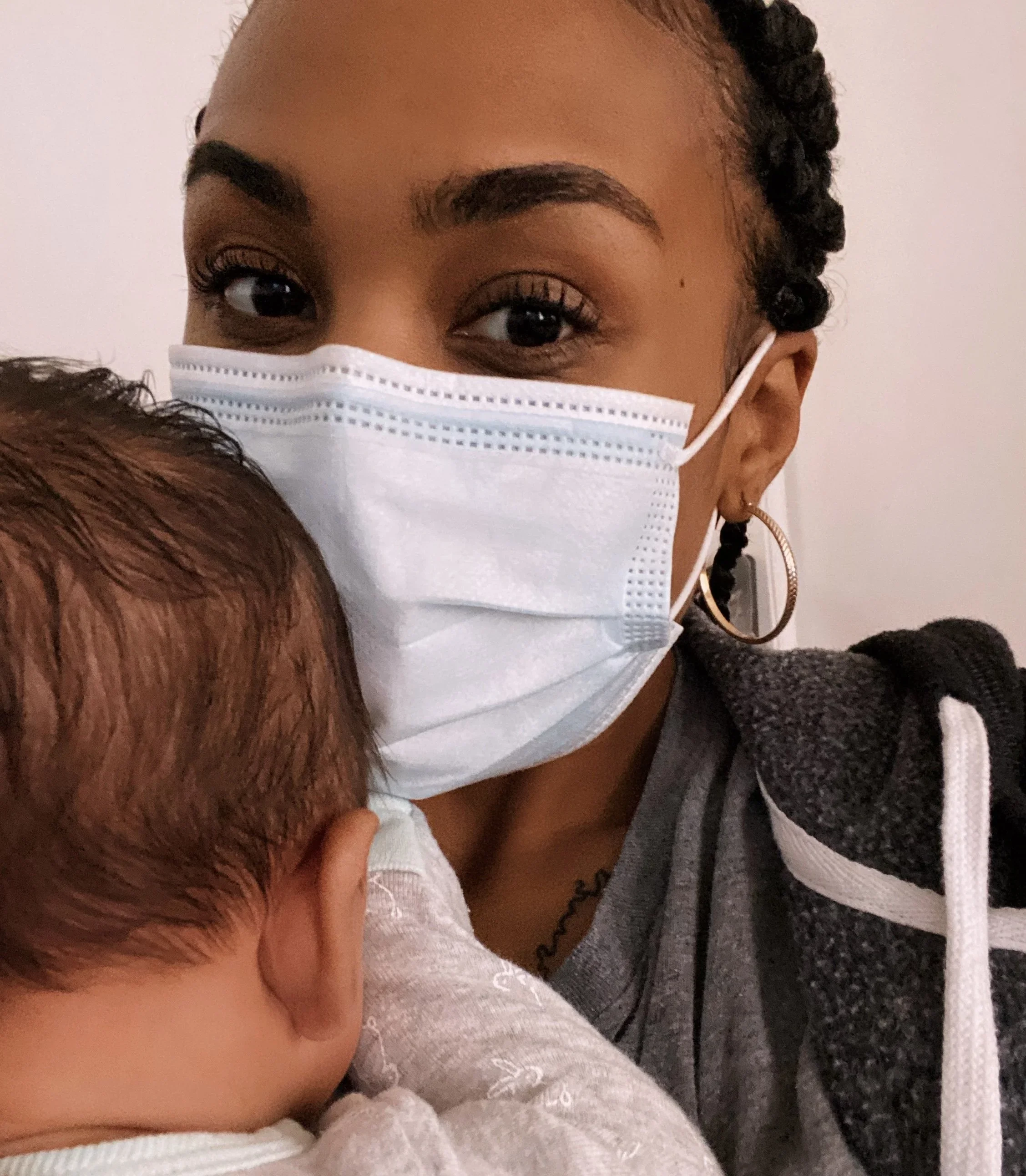 A woman wearing a face mask holding a young child. The woman has dark braided hair, hoop earrings, and is dressed in a casual hoodie. The child's head is resting on her shoulder, showing brown hair and part of a gray shirt.