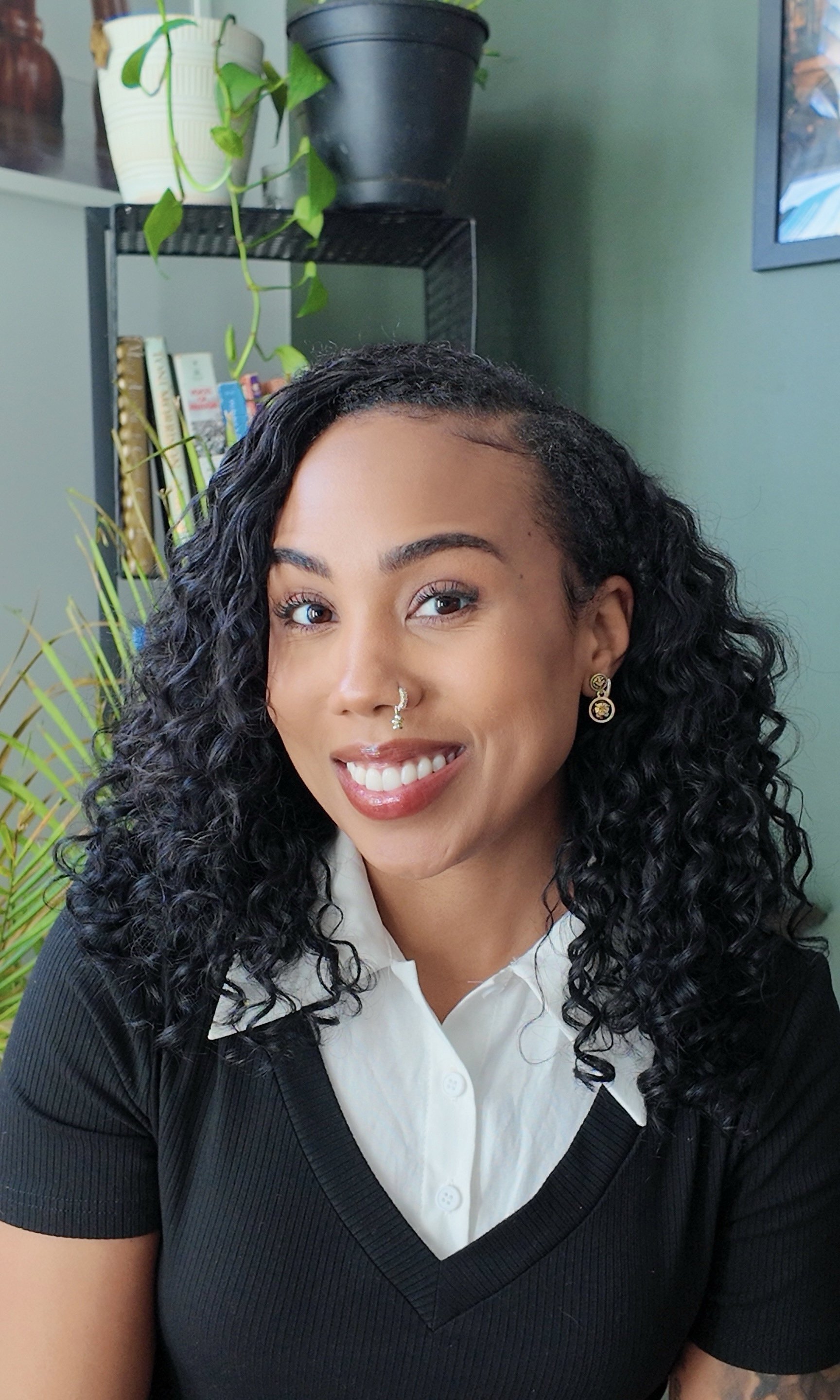 A woman with curly black hair, smiling, wearing a nose ring, earrings, a white collared shirt, and a black sweater, sitting in front of green walls and a bookshelf with plants and books.