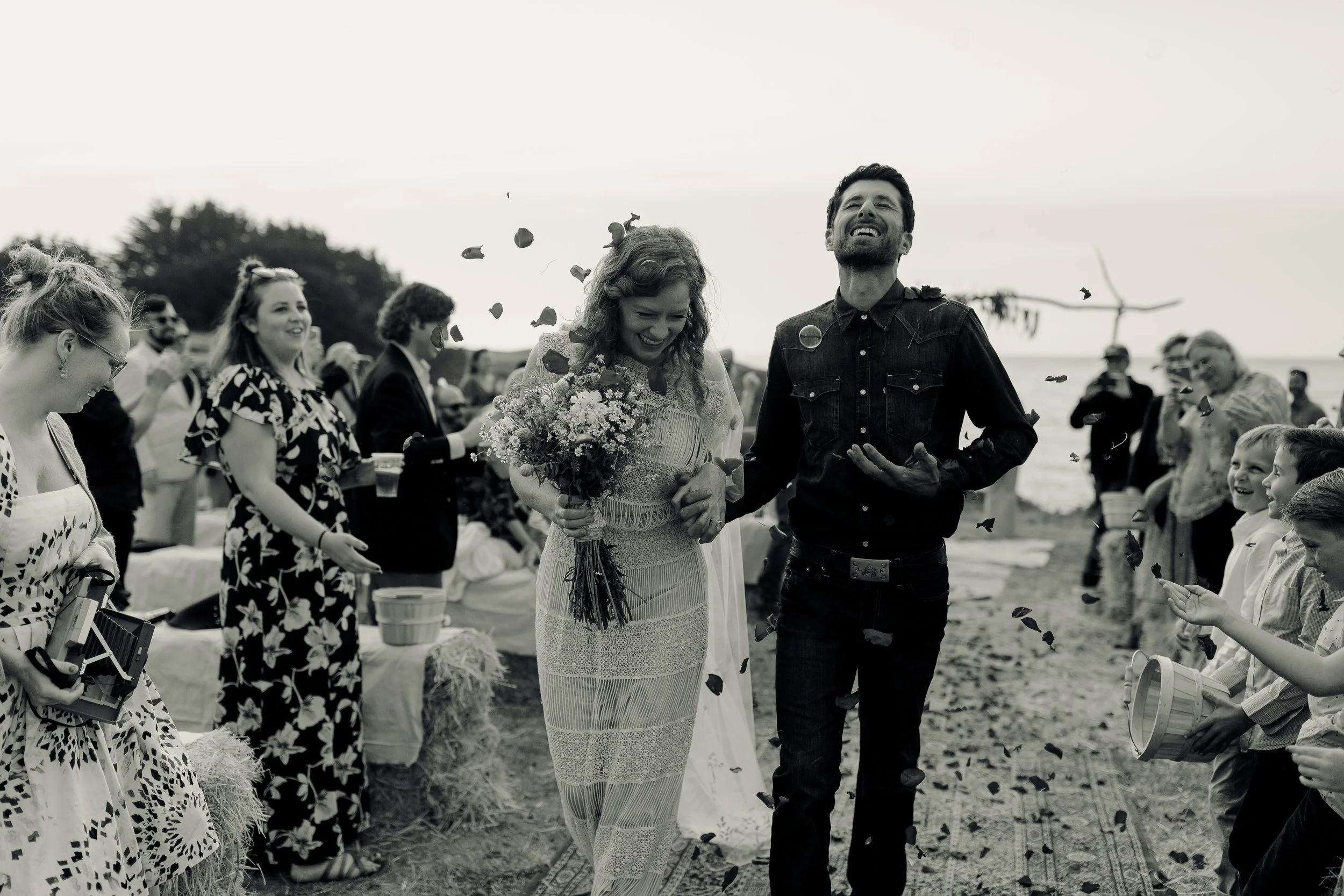 Newlyweds walking down an outdoor aisle as guests toss petals beside the ocean.