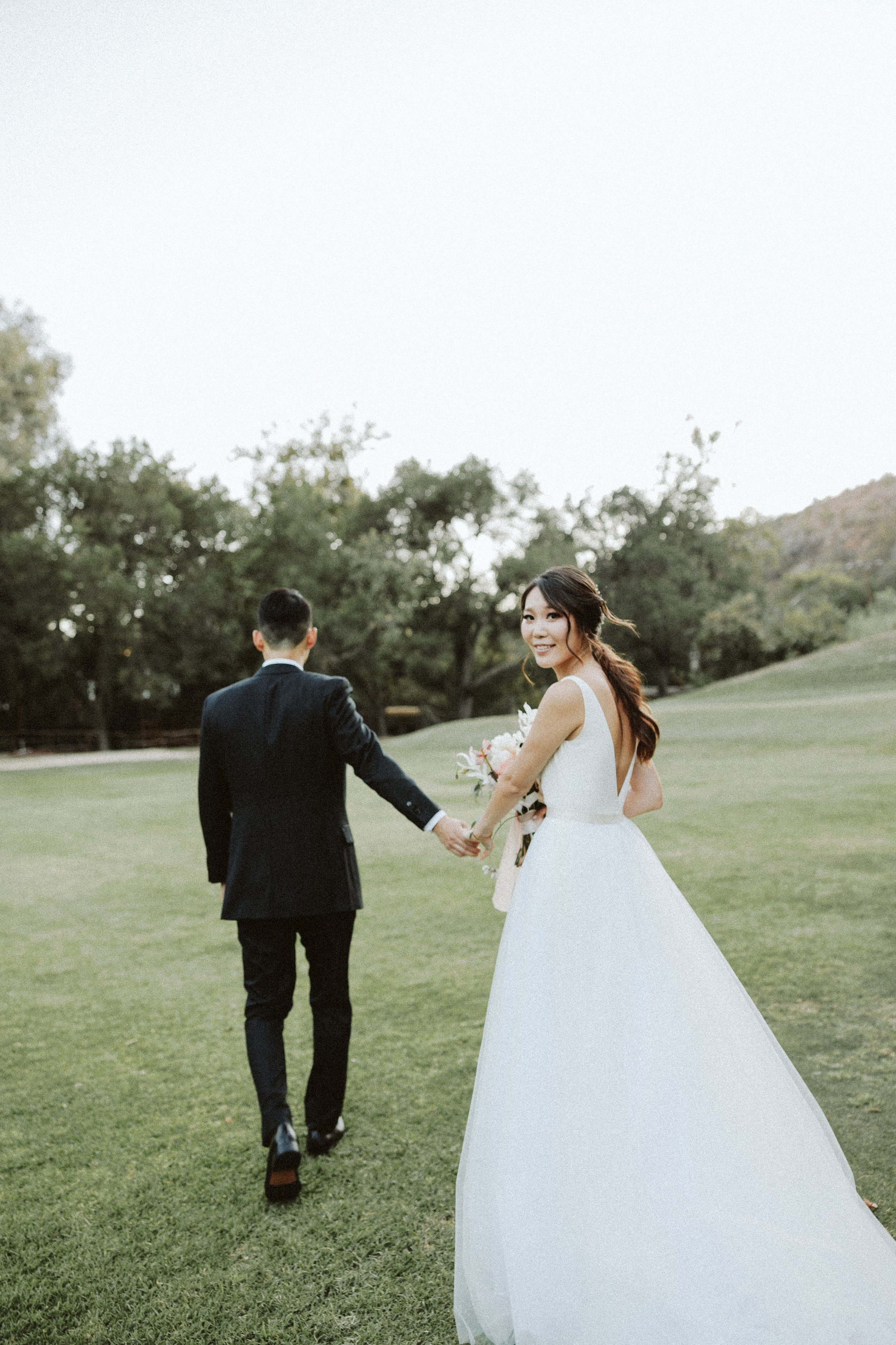 Newlyweds walking hand in hand across a lawn as the bride looks back over her shoulder.