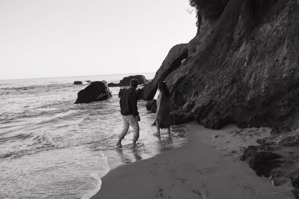 Black and white photograph of a couple walking through shallow surf beside a rocky coastal cliff.
