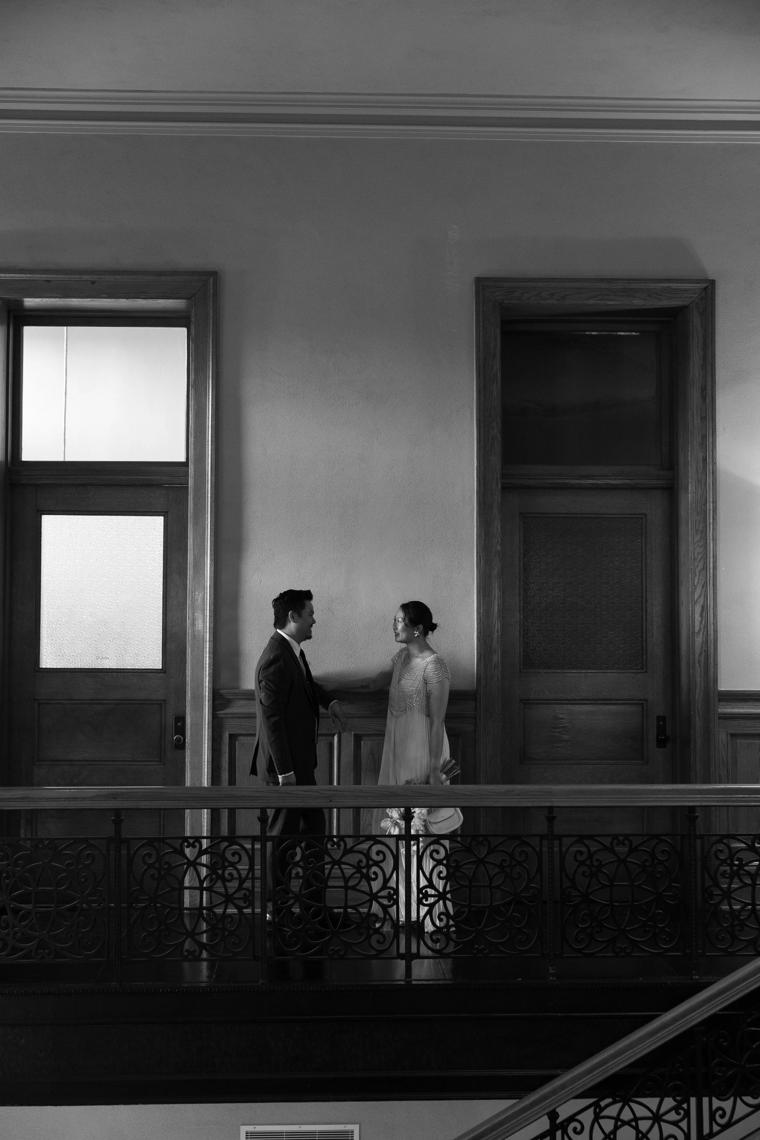 Black and white wedding portrait of a couple facing each other on an interior balcony beside tall windows.