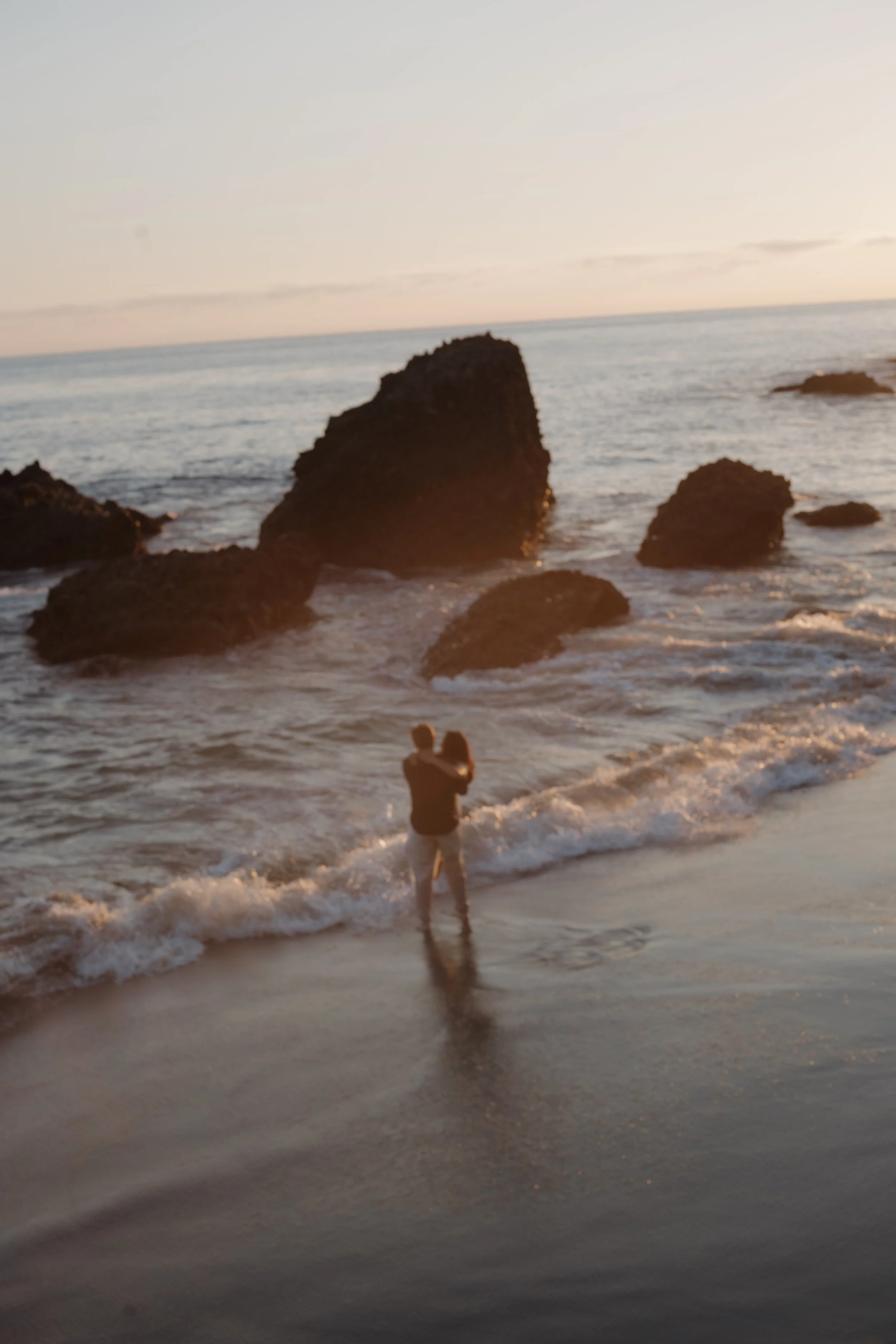 Couple standing together in the surf at sunset, with large rocks rising from the ocean behind them.