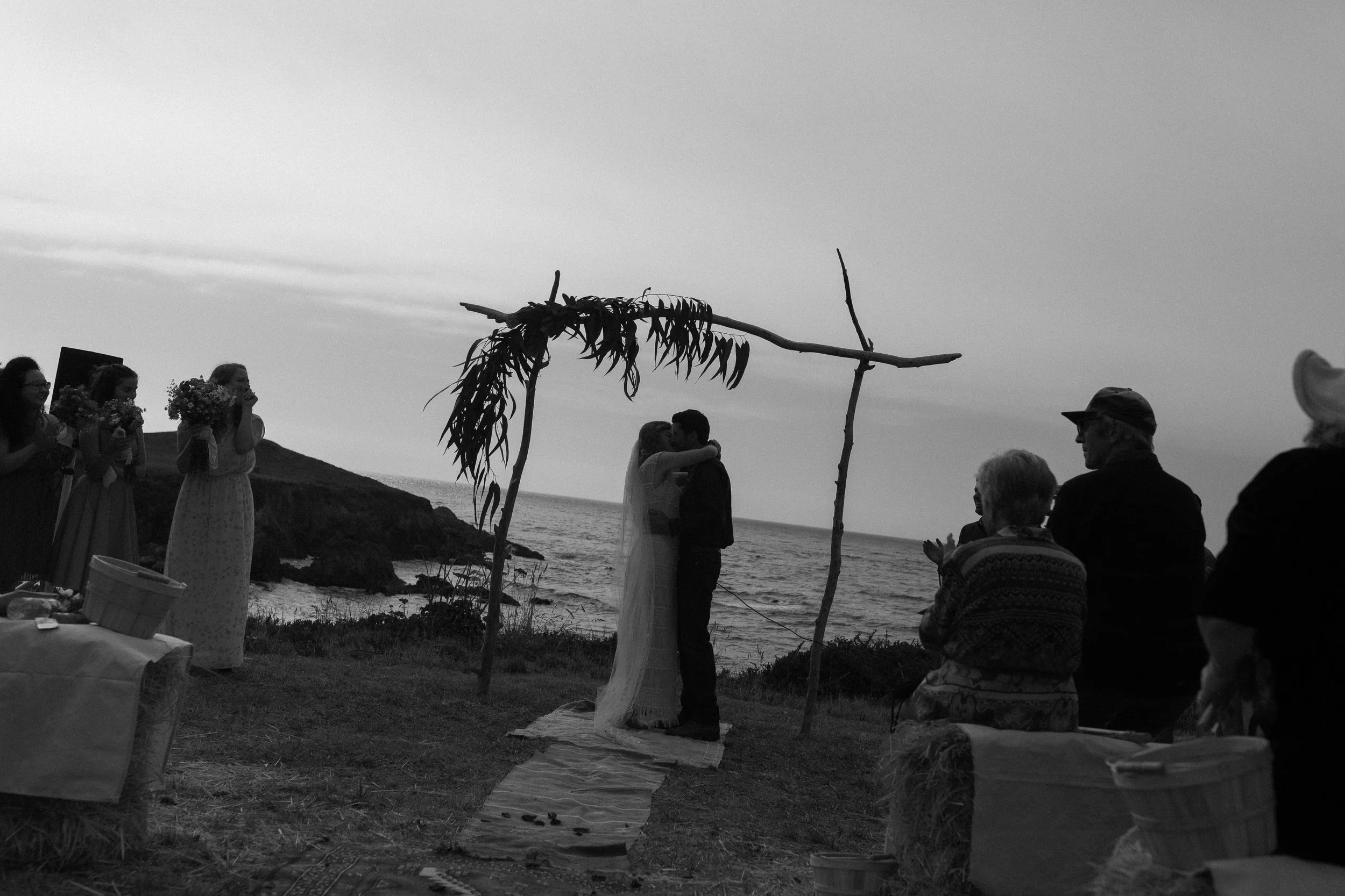 Black and white wedding photograph of a couple kissing beneath a wooden arch overlooking the ocean.