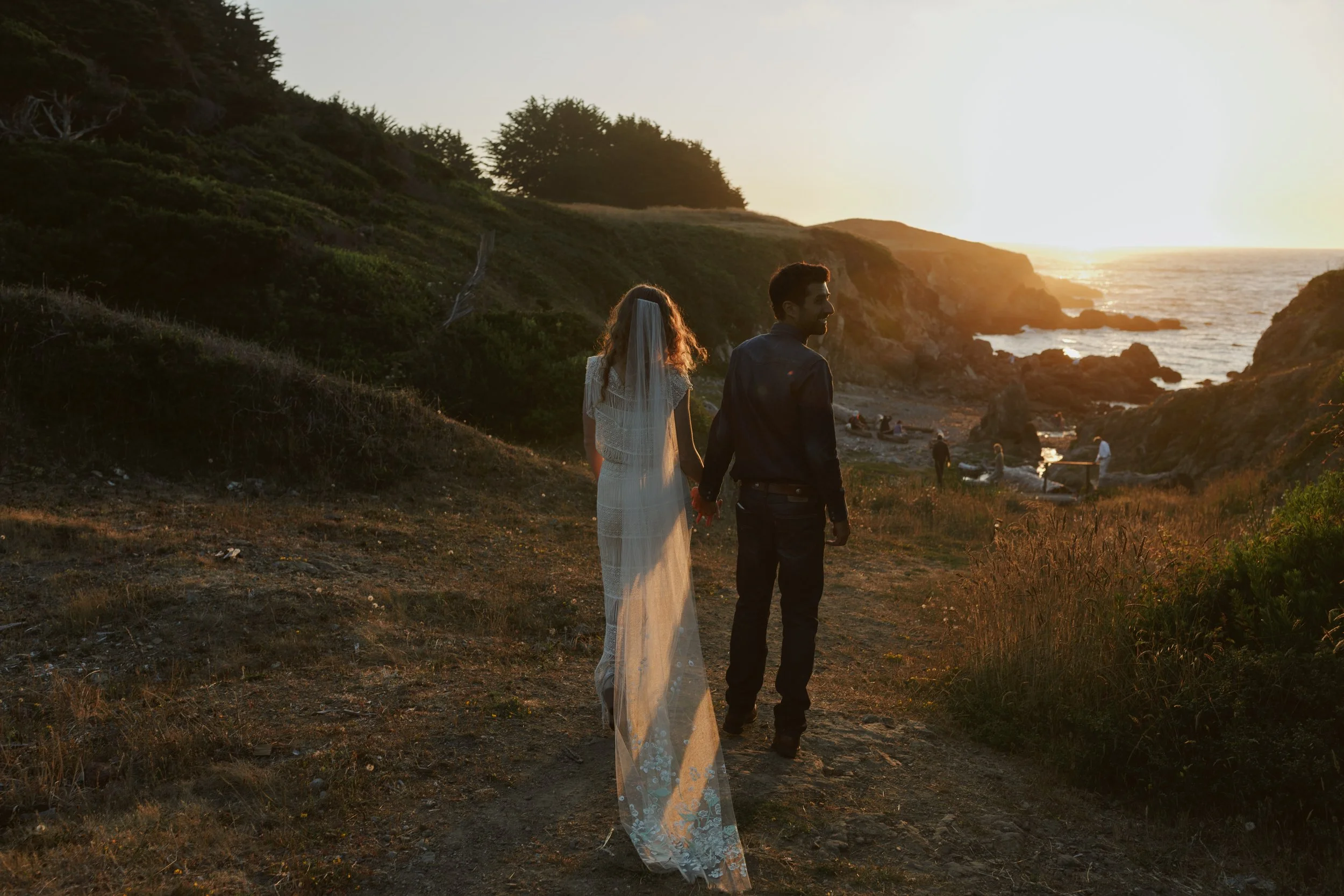 Newlyweds holding hands on a coastal path at sunset, with the bride’s veil trailing behind her.