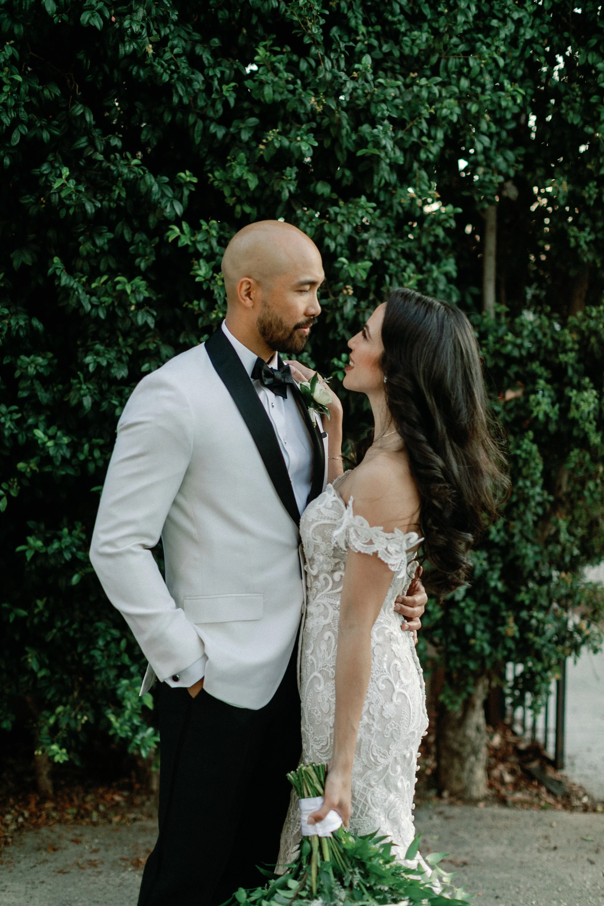 Bride and groom facing each other in a garden, framed by green foliage.