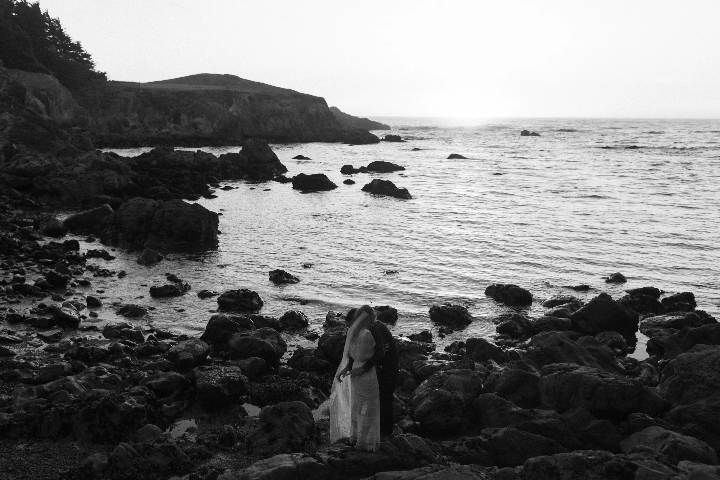Black and white portrait of newlyweds among rocks beside the ocean.