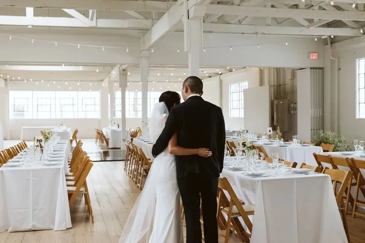 Bride and groom standing arm in arm in a sunlit reception room with white tablecloths and wooden chairs.