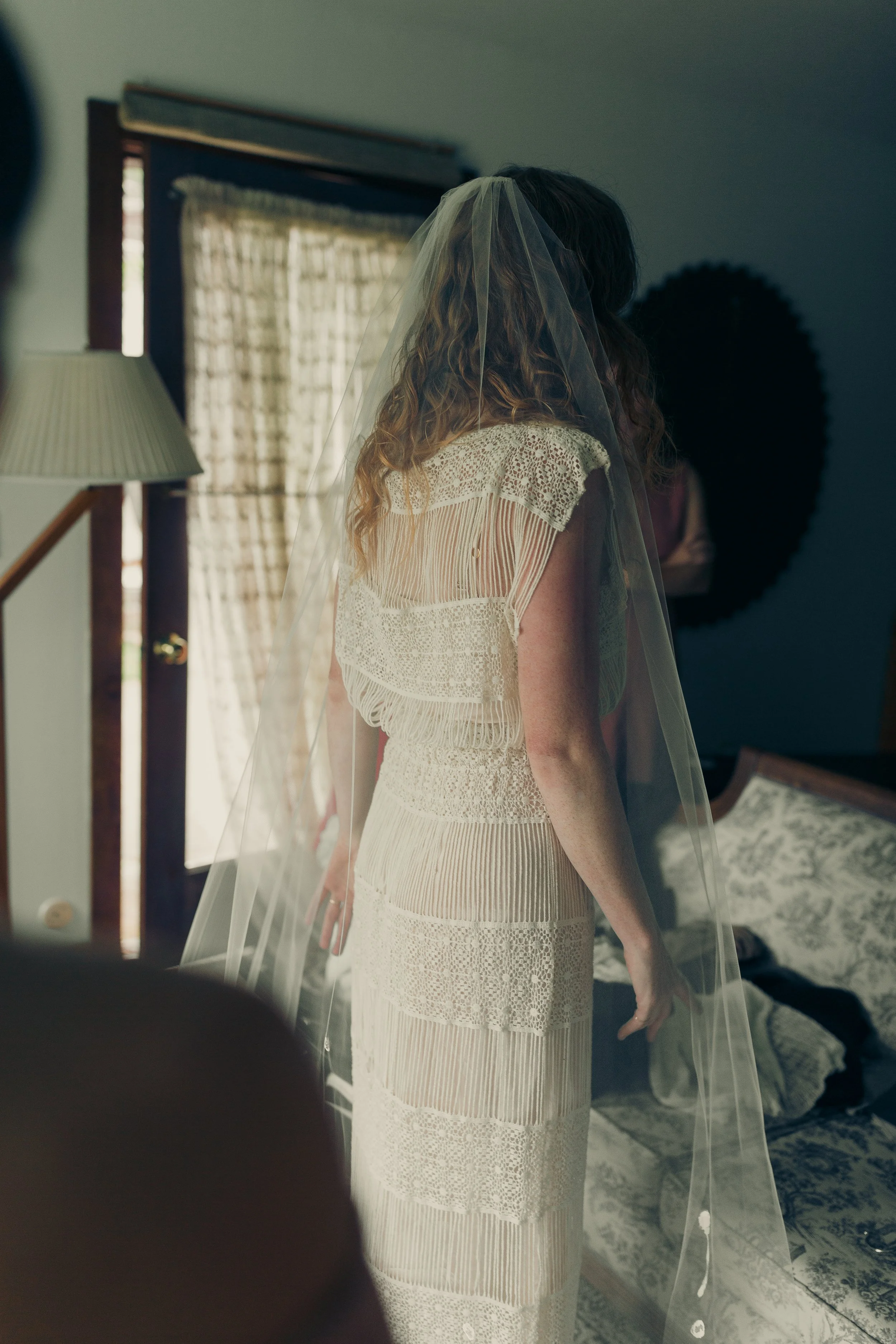 Bride in a lace wedding dress and veil standing beside a window in a softly lit room.
