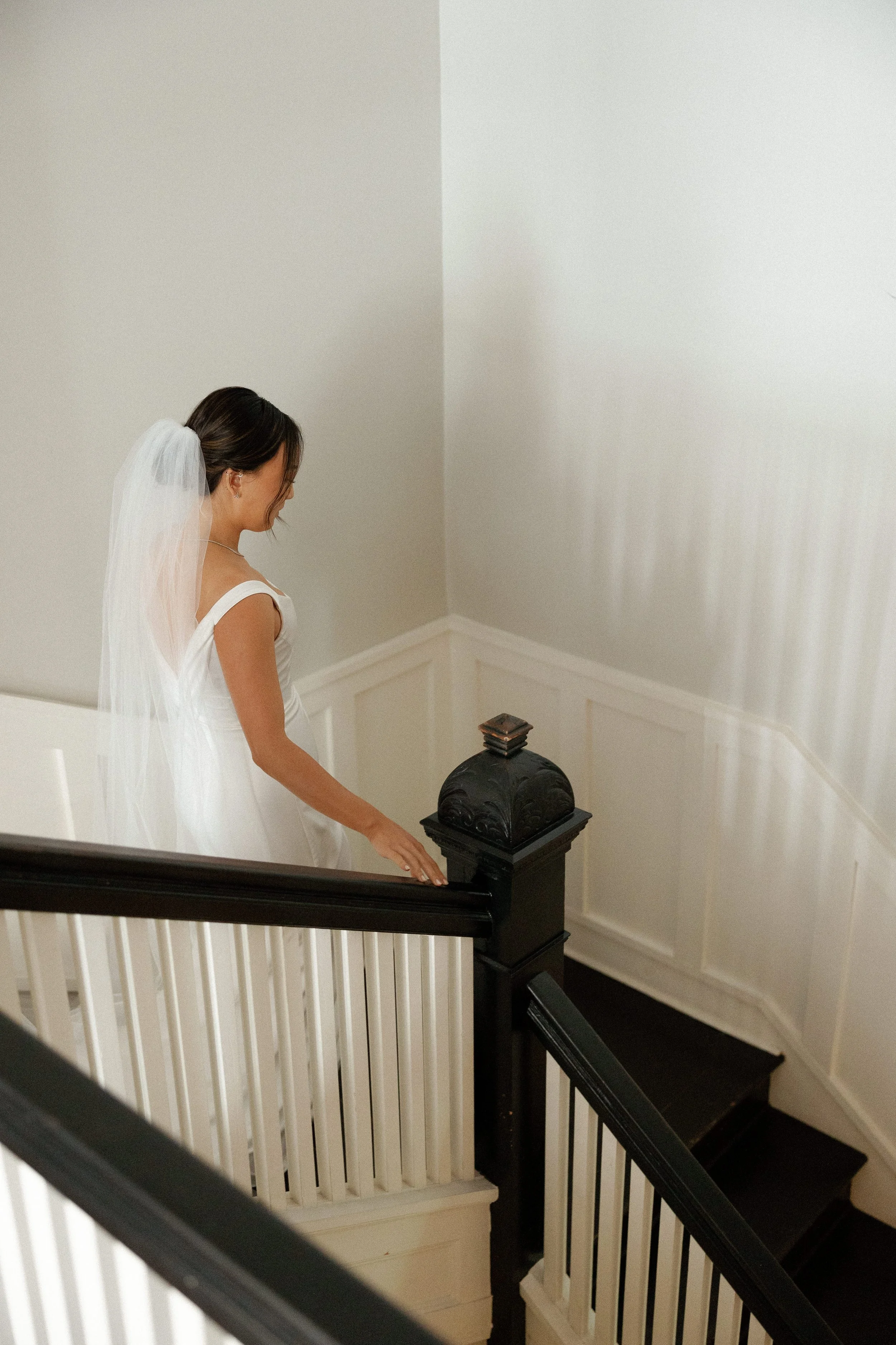 Bride in a white wedding dress and veil descending a staircase beside a dark wooden handrail.