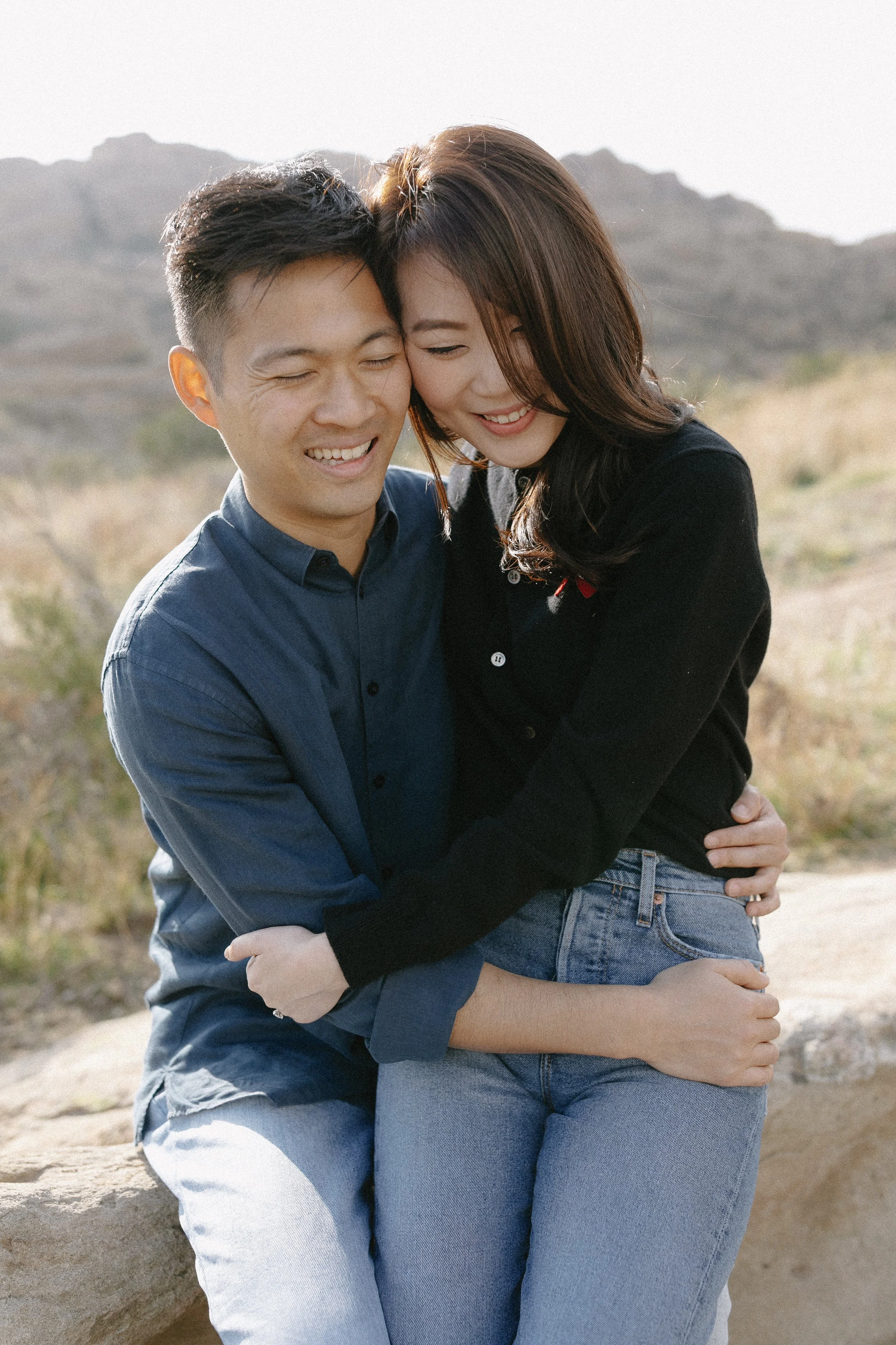 Couple embracing and smiling during an outdoor engagement session with rocky hills behind them.