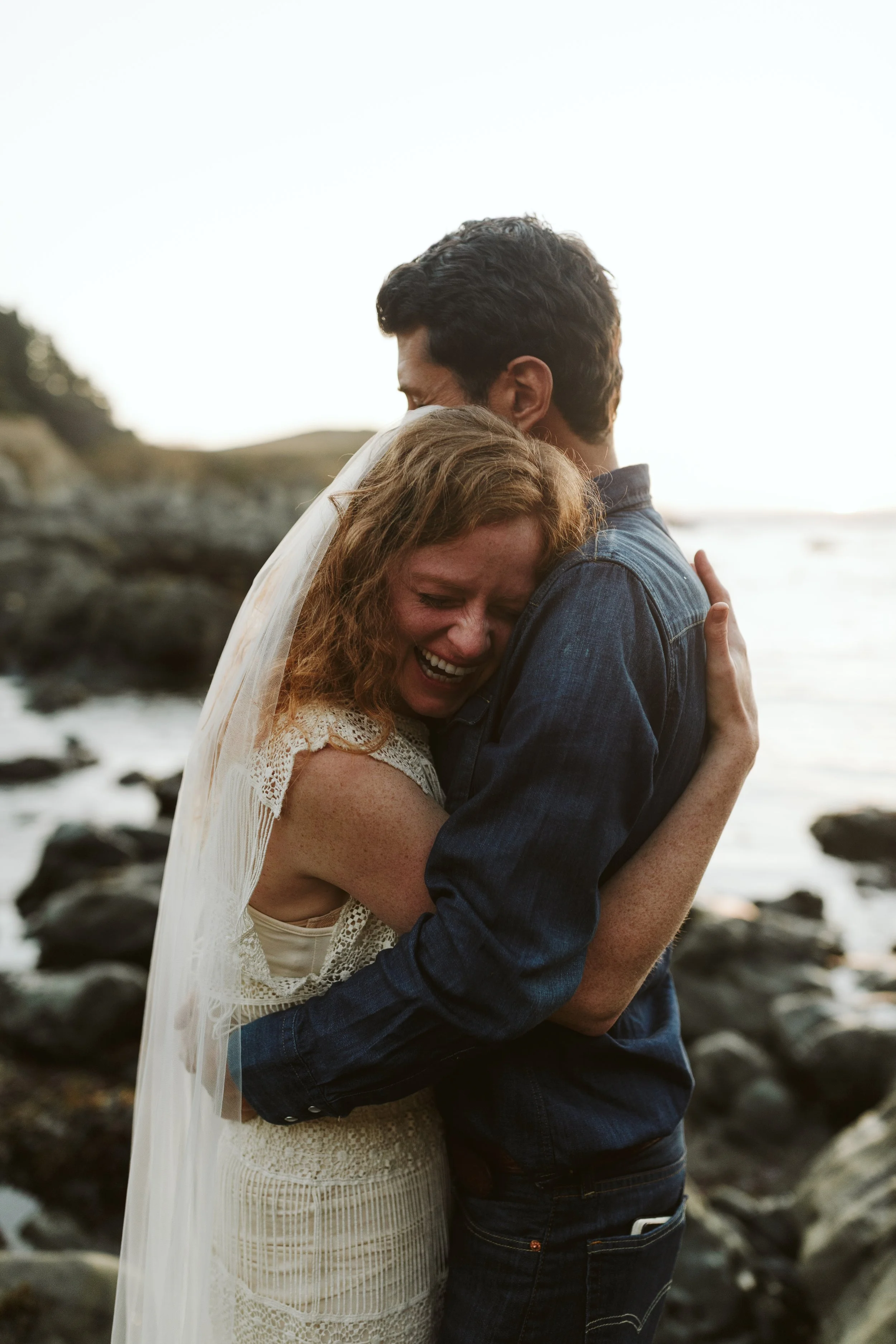 Bride laughing as she leans into the groom during a seaside embrace.
