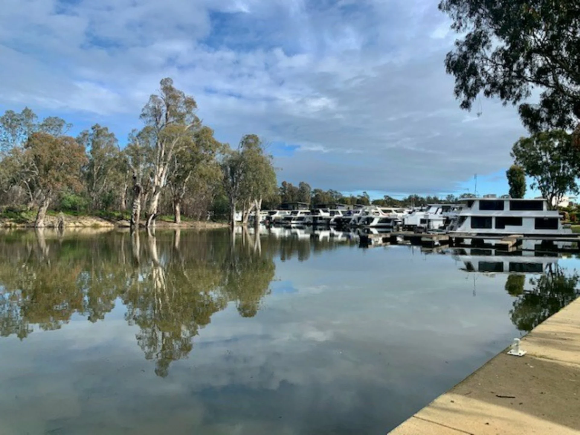 Calm water reflecting trees and docked houseboats along a shoreline under a partly cloudy sky.