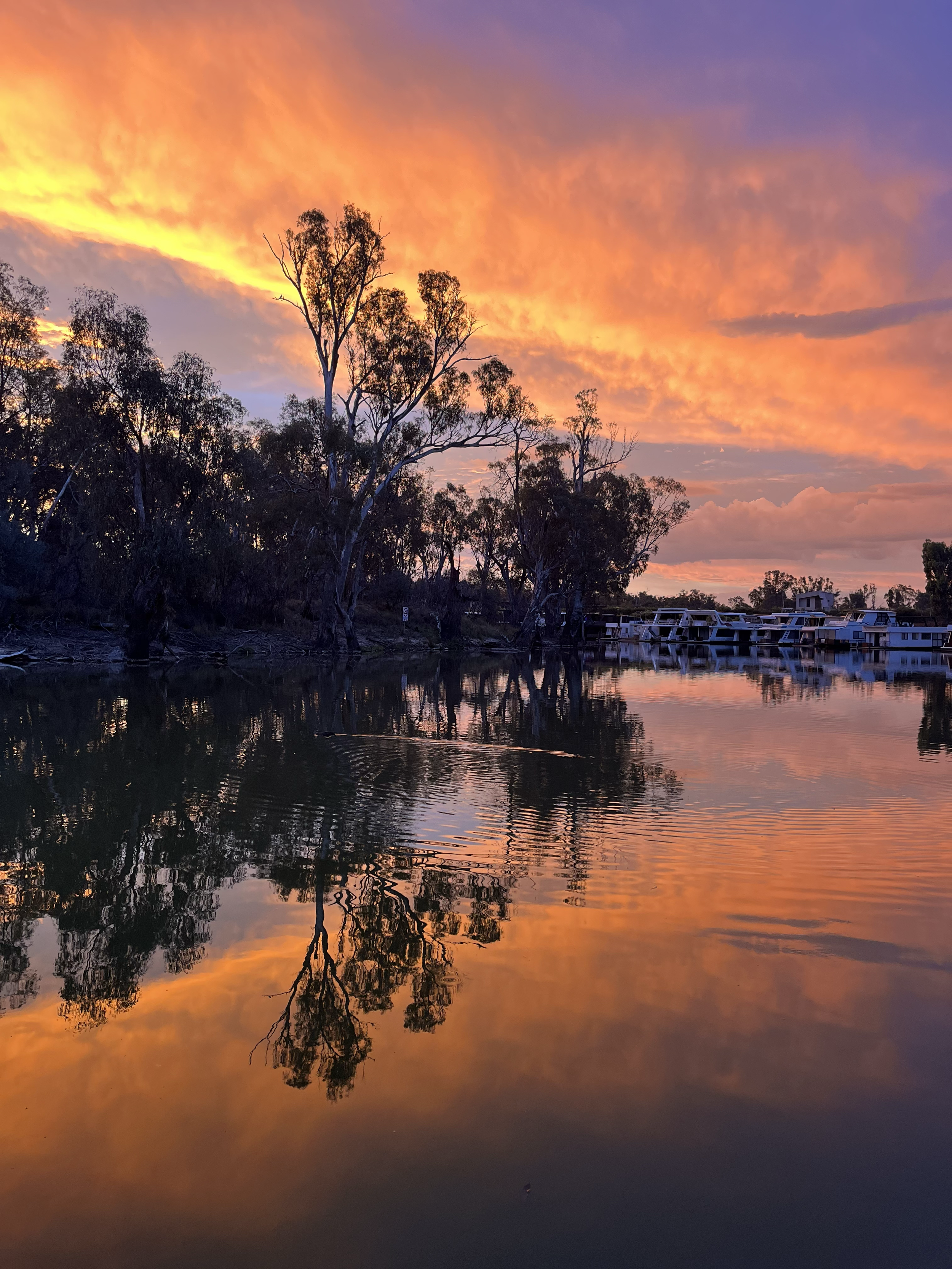 A vibrant sunset over a calm river, with silhouetted trees and boats along the shore, reflecting the colorful sky in the water.