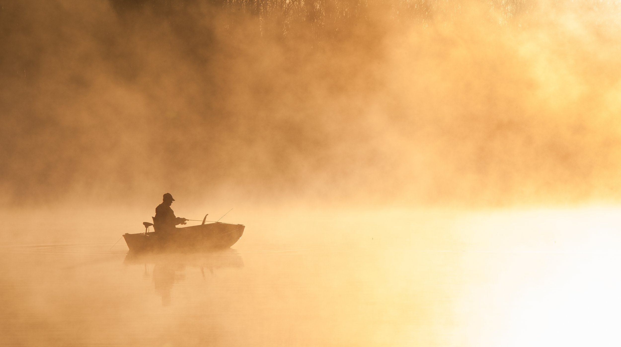 Silhouette of a person fishing from a small boat on calm water with a foggy or misty background, golden hue.