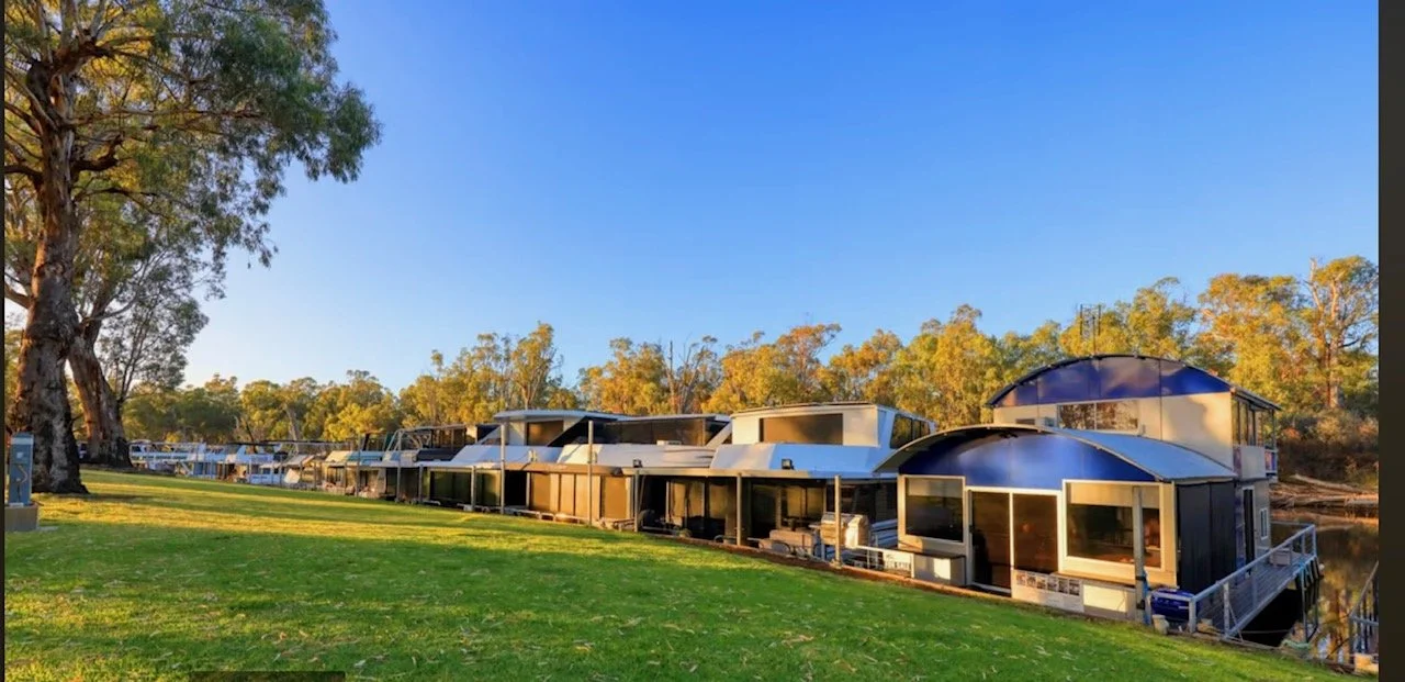 Line of houseboats docked along a grassy shoreline with trees and a blue sky in the background.