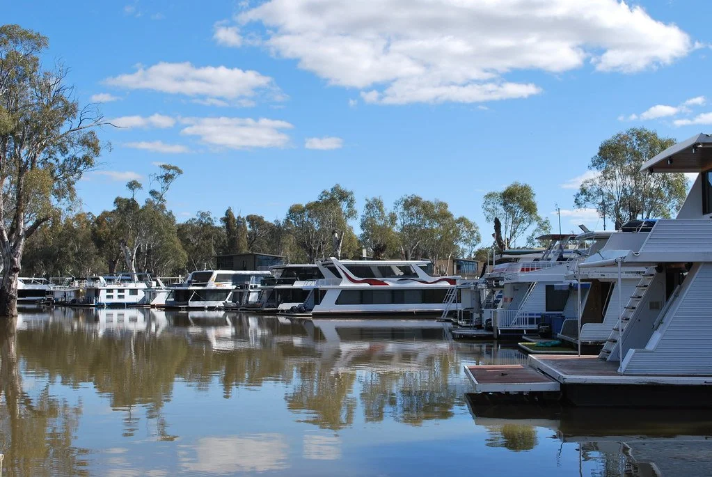 Marina with several boats docked along a calm waterway, with trees and houses in the background and a partly cloudy sky above.