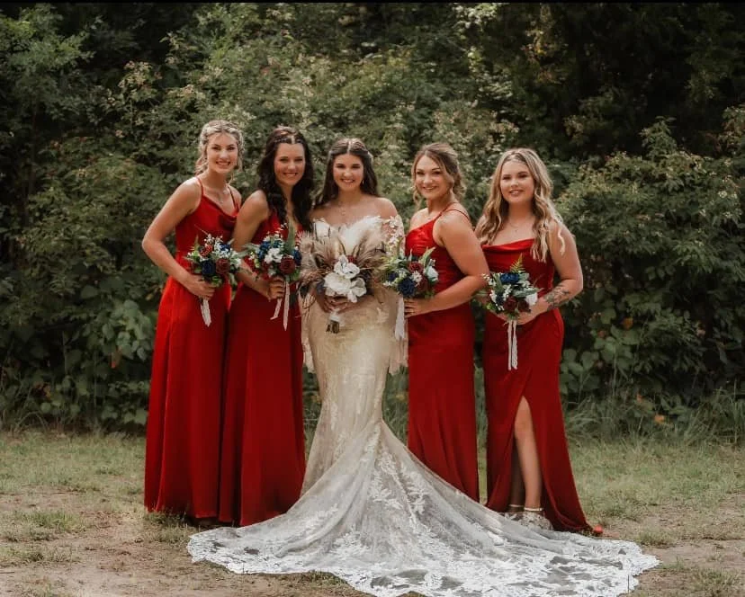 A bride in a white wedding gown surrounded by four bridesmaids in red dresses, standing outdoors in front of green foliage, holding bouquets.