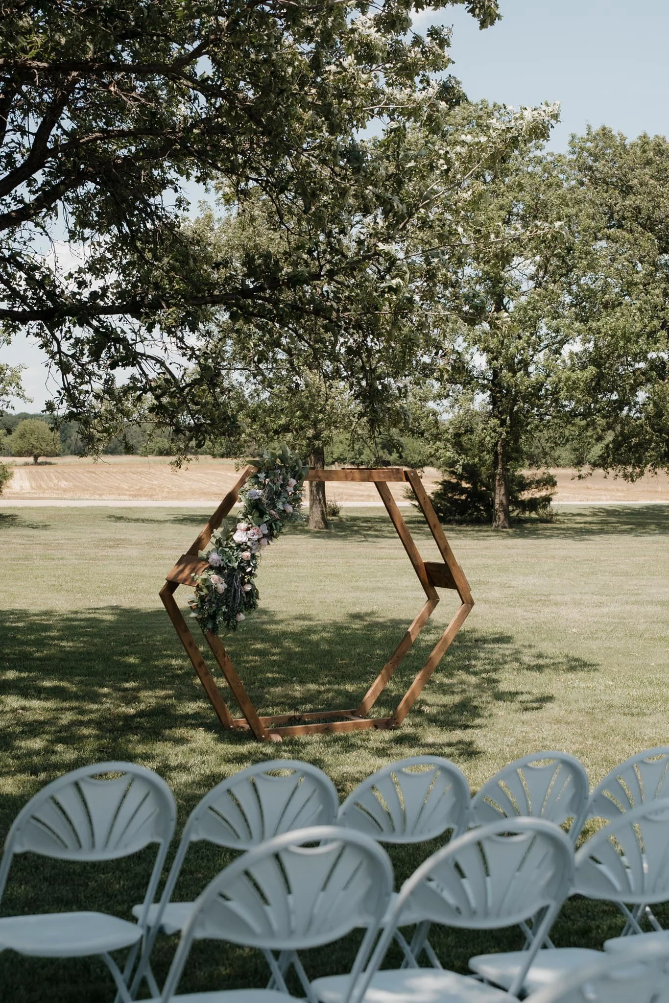 Outdoor wedding ceremony setup with a wooden hexagonal arch decorated with flowers, under a large leafy tree, with rows of white chairs facing the arch.