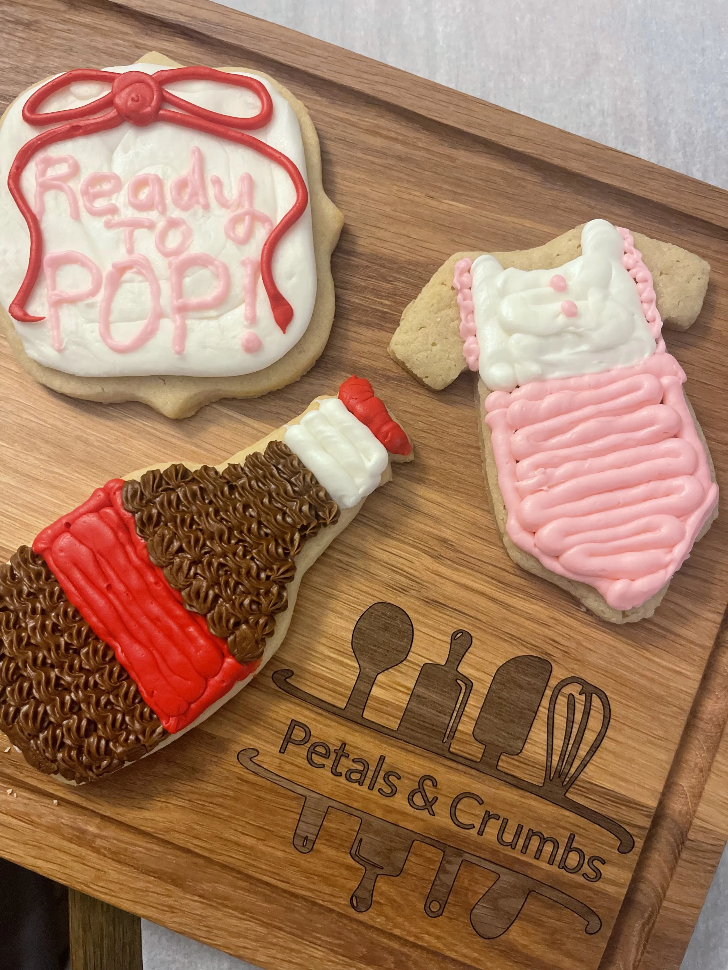 Decorated sugar cookies on a wooden tray with the logo "Petals & Crumbs"; cookies include a bag with "Ready to Pop!" written, a baby bottle, and a bottle of nail polish with a red top.