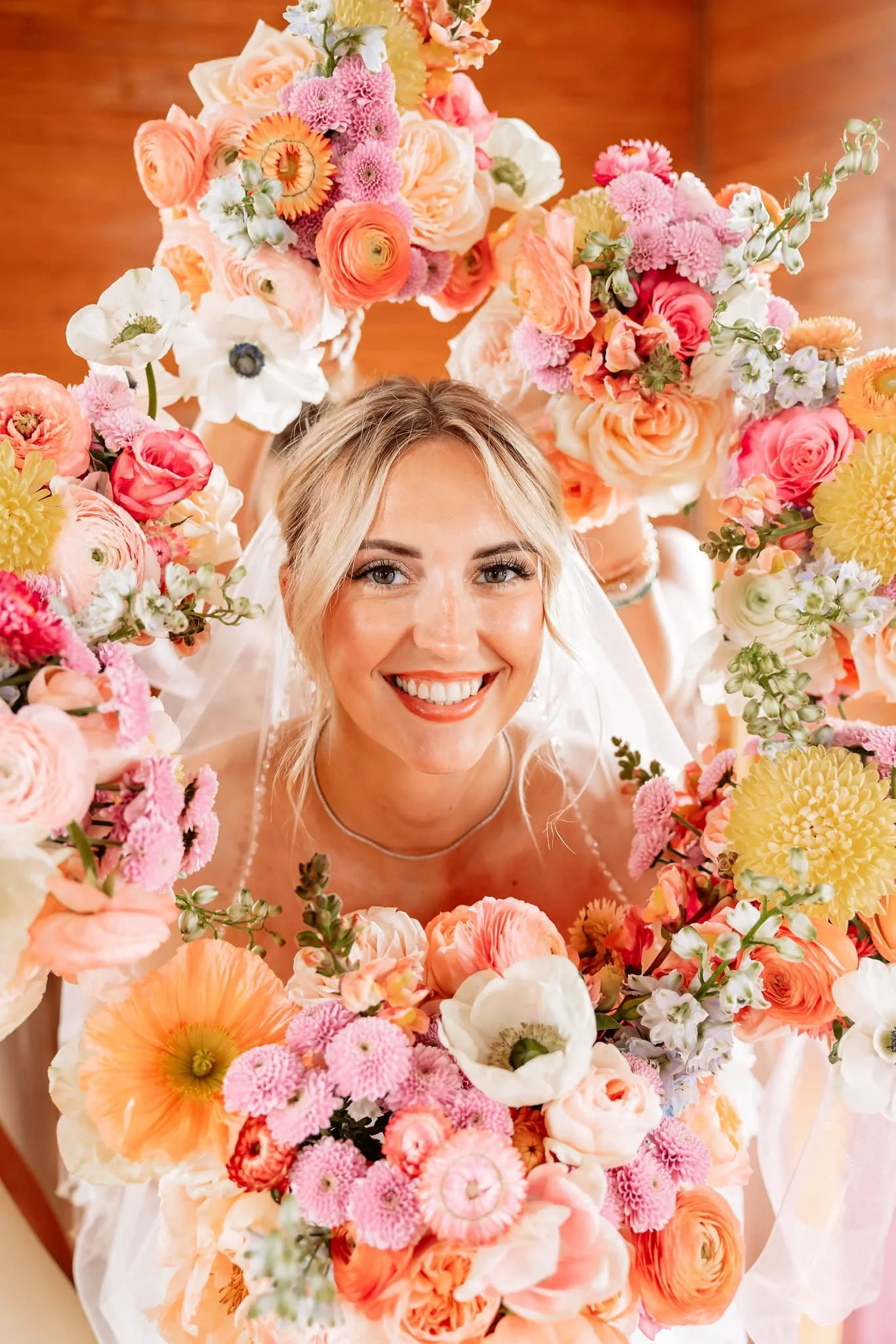 A smiling bride surrounded by colorful flowers, including roses, ranunculus, anemones, and other blossoms, arranged in a floral arch.