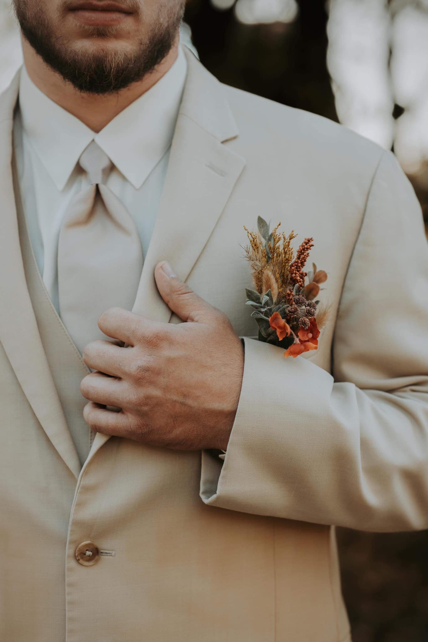 Groom wearing a beige suit with a flower boutonniere, holding the lapel of his jacket with his right hand.