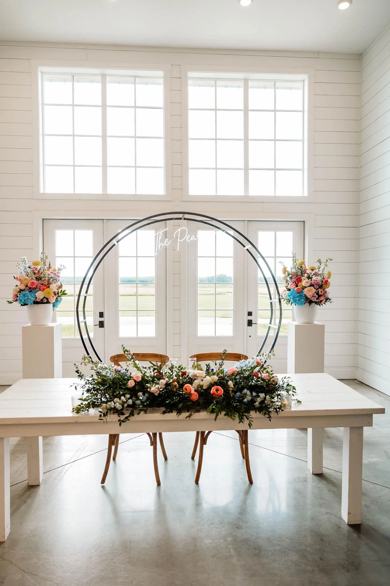 A decorated table with floral arrangements and a horseshoe-shaped backdrop inside a bright, white interior with large windows.
