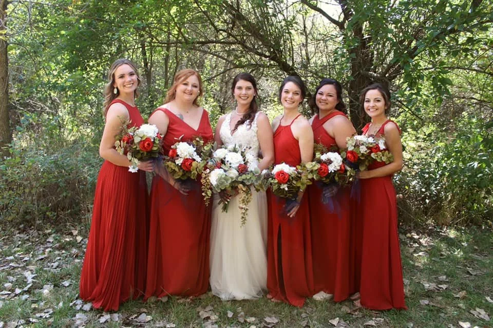 Group of six women in long red dresses holding bouquets of flowers, standing outdoors in a wooded area.