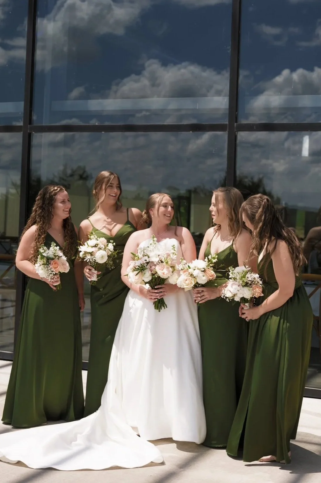 Bride in white wedding dress with five bridesmaids in green dresses, holding bouquets of flowers, standing outside in front of a glass building reflecting a cloudy sky.