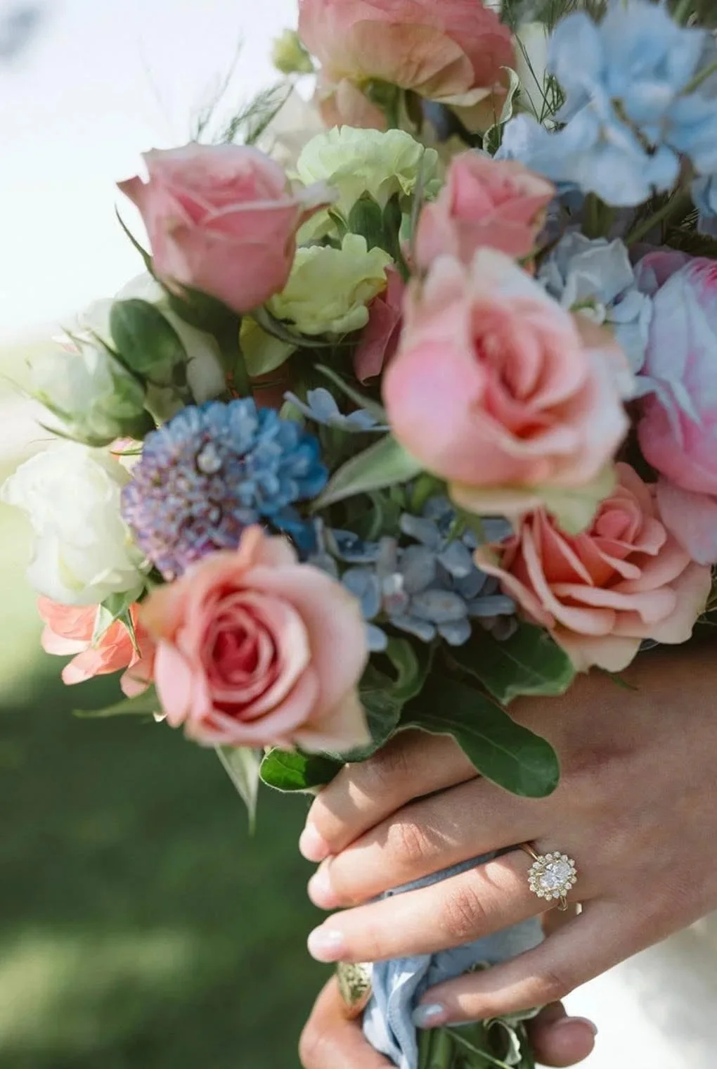 A bouquet of pink, white, green, and blue flowers held by a person wearing a ring with a floral design on their finger.