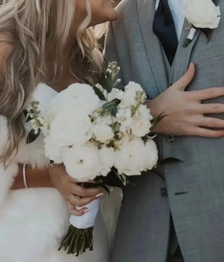 Close-up of a bride holding a white flower bouquet, with a groom partially visible wearing a gray suit and black tie, at a wedding ceremony.