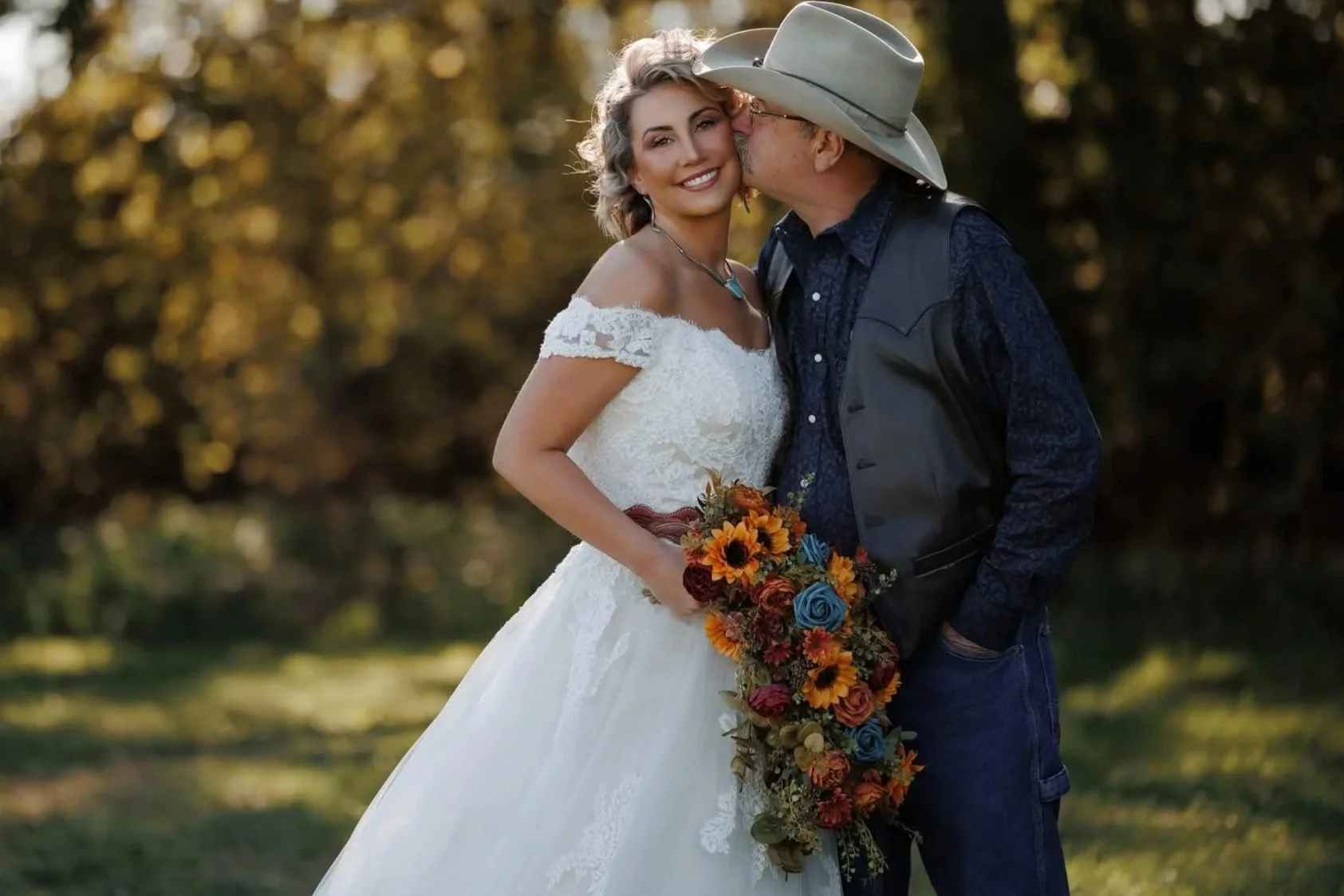 A bride in a white wedding dress with lace details is being kissed on the cheek by an older man wearing a cowboy hat, vest, and a dark shirt. The bride is holding a colorful bouquet of flowers, and they are standing outdoors during the daytime with t
