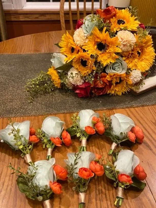 A bouquet of mixed flowers including sunflowers, roses, and carnations on a wooden table, with several boutonnières made of white roses and orange buds arranged in front.