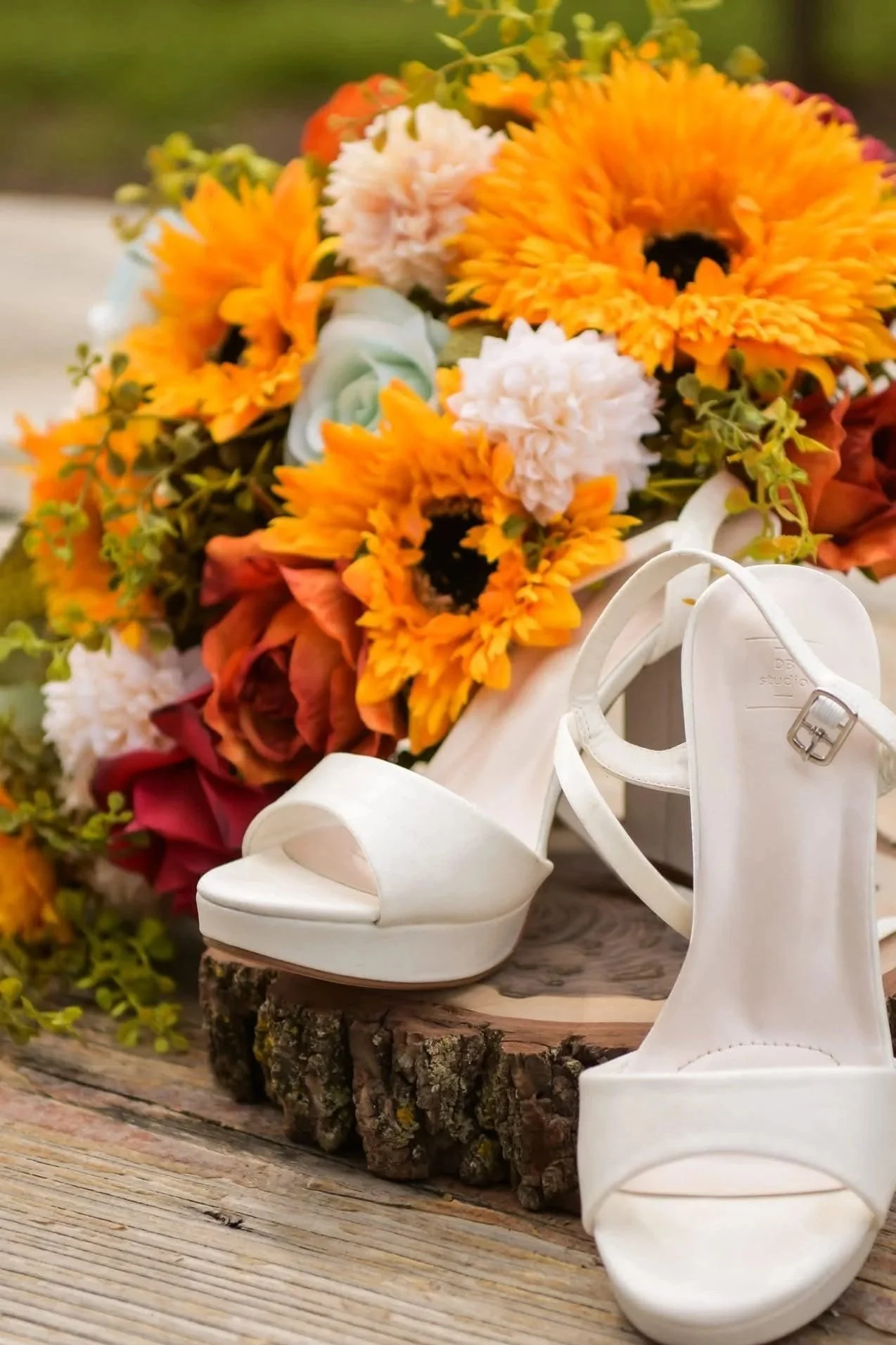 A pair of white high-heeled sandals placed on a wooden log, with a colorful bouquet of sunflowers, roses, and other flowers in the background.