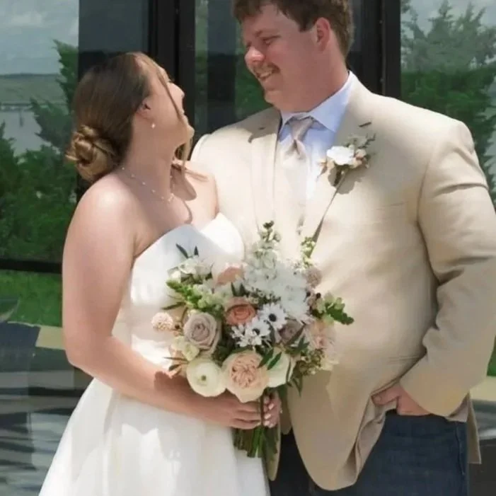 A bride and groom on their wedding day, smiling at each other, with the bride holding a bouquet of flowers and dressed in a white wedding gown, and the groom wearing a light-colored suit.