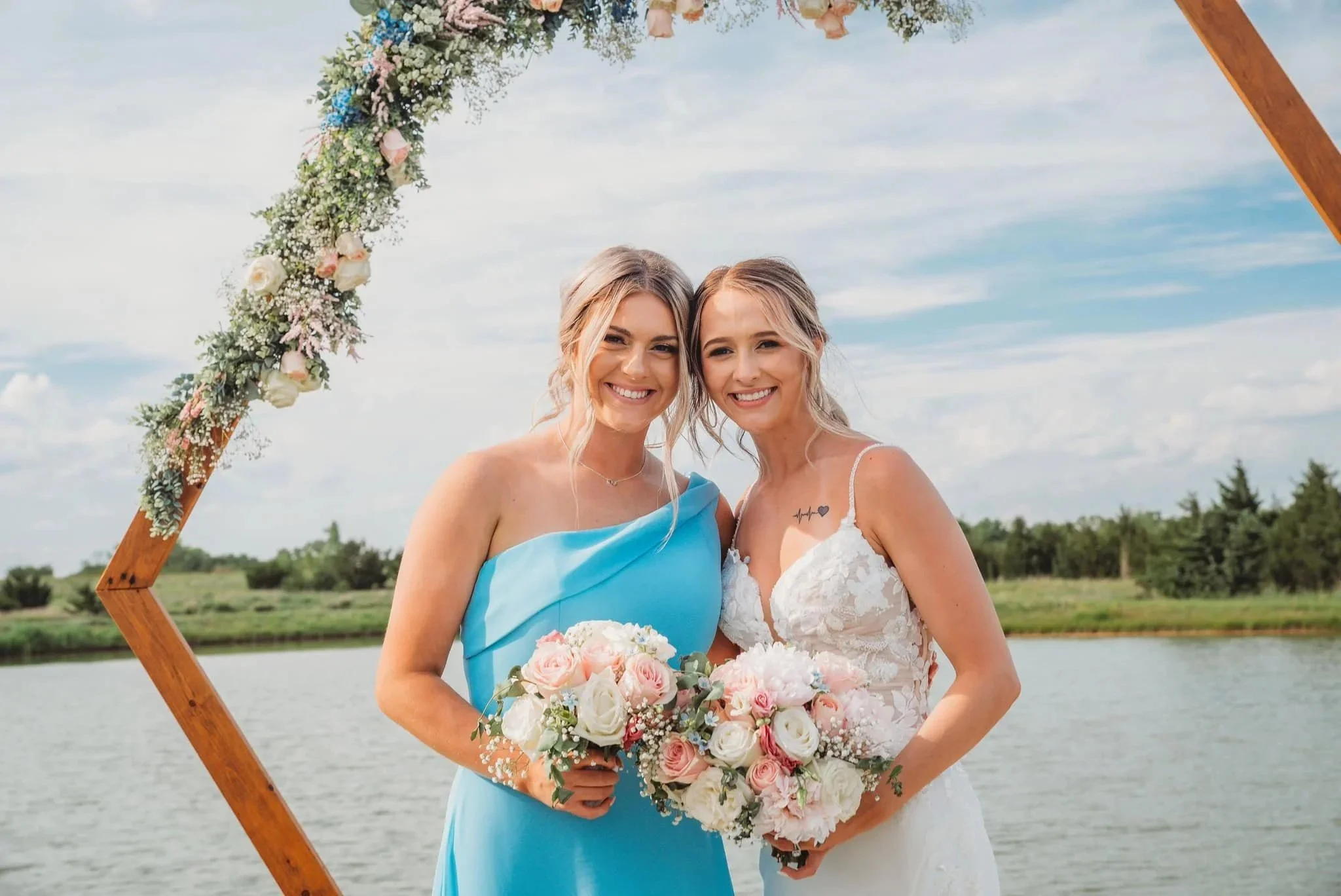 Two women in wedding attire standing outdoors near a lake, holding bouquets of flowers, smiling, with a wooden wedding arch decorated with flowers behind them.