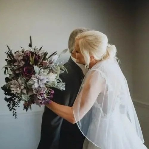 An elderly couple hugging, she is holding a bouquet of flowers in a wedding dress, he is dressed in a suit.
