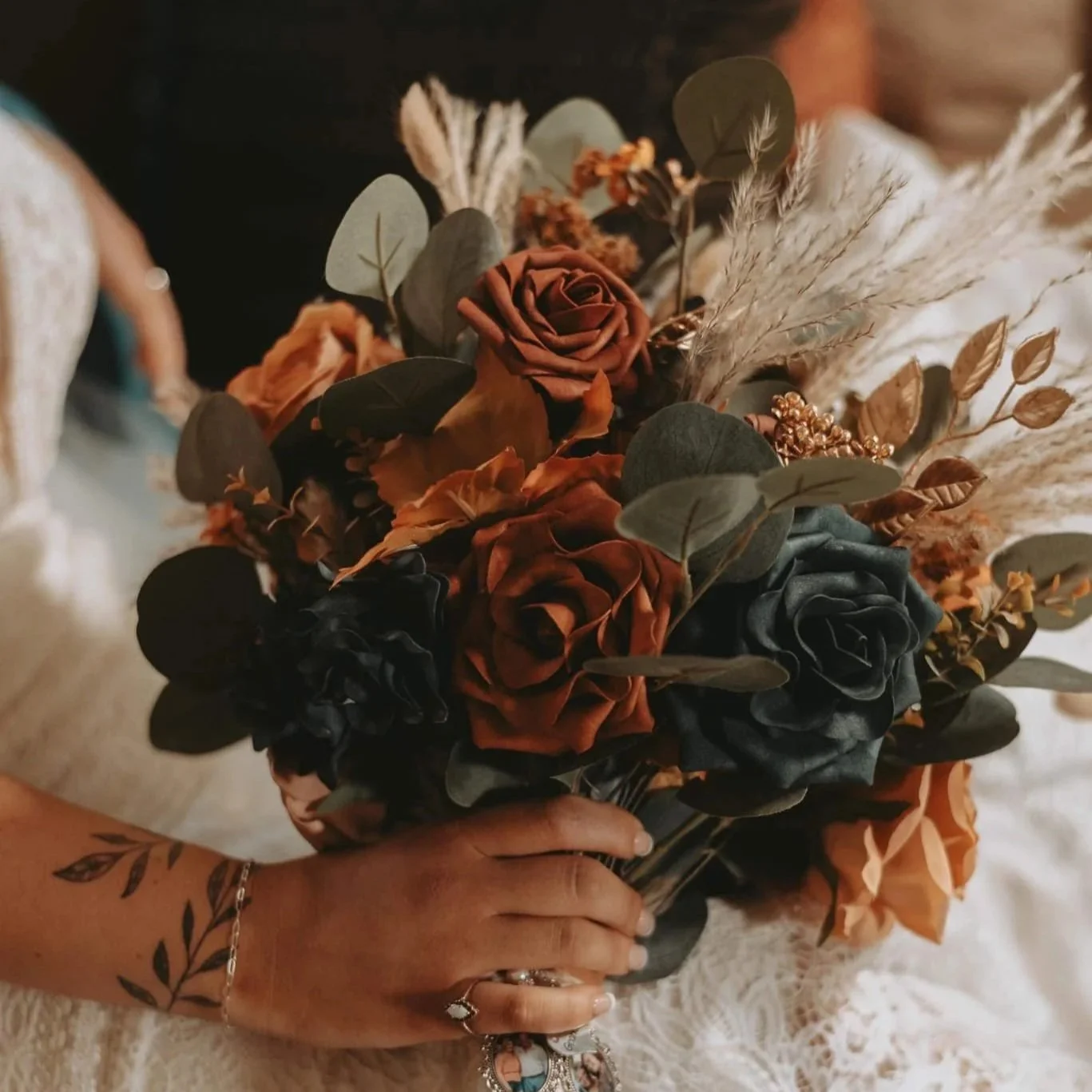 A person holding a bouquet of flowers consisting of brown, turquoise and orange roses, along with green foliage and beige dried grasses.
