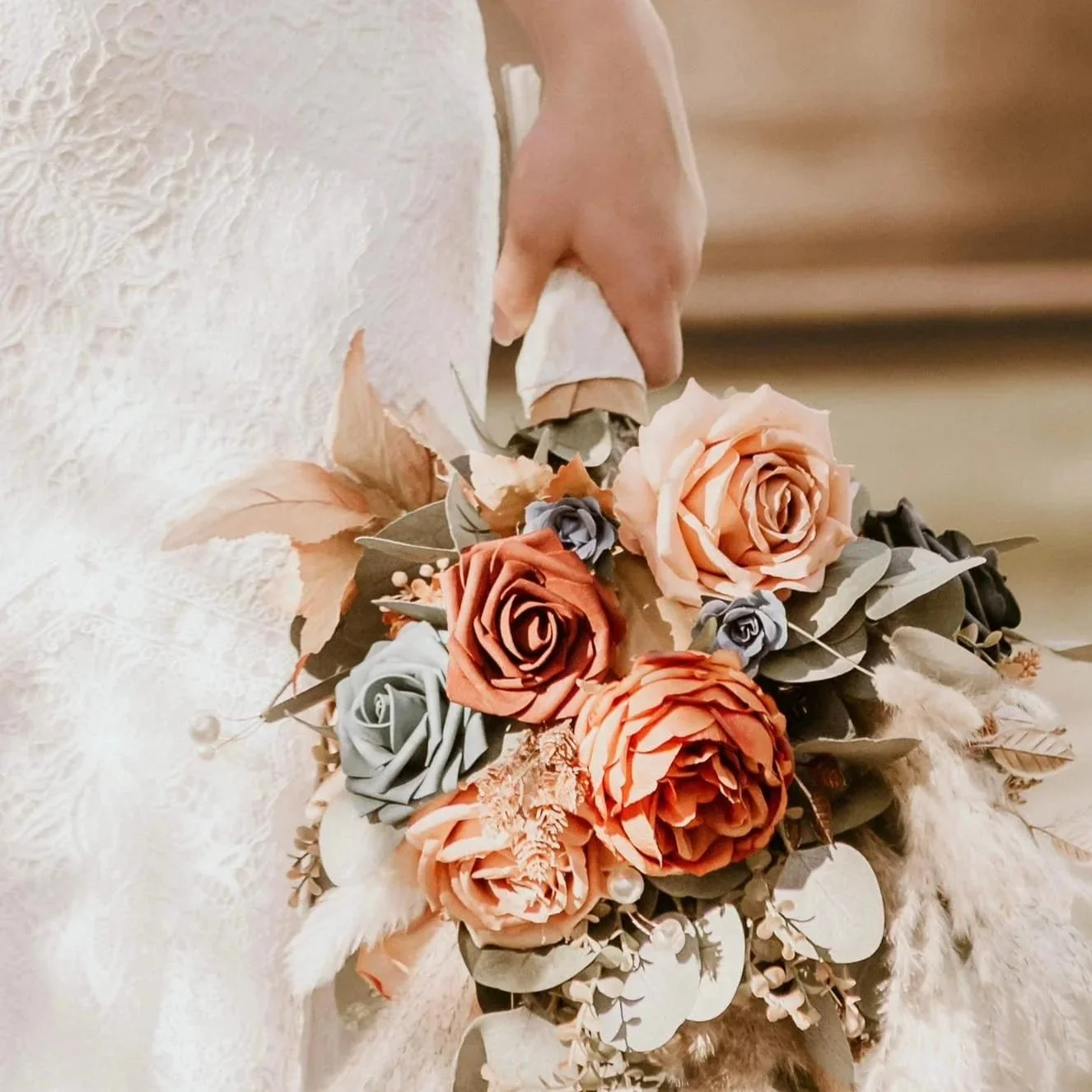 A person holding a bouquet of peach, gray, and coral roses with various foliage and decorative elements.